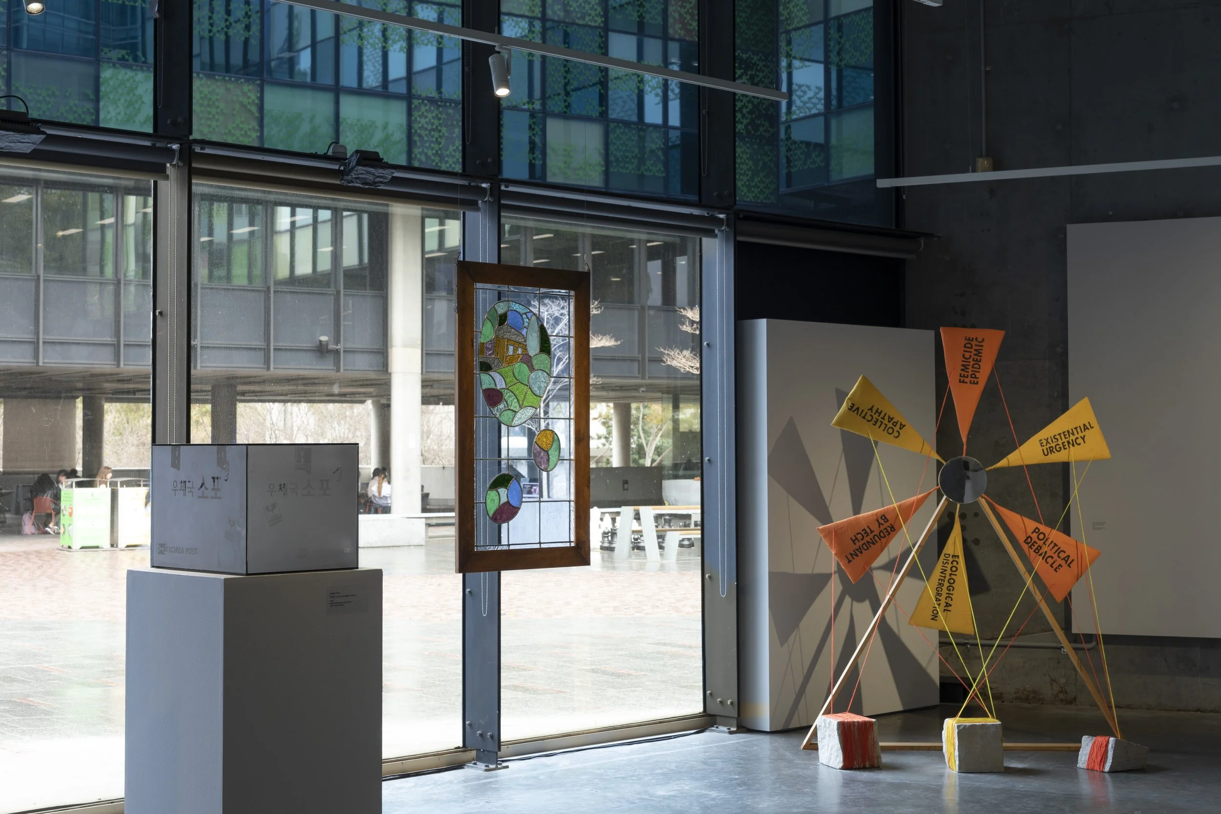 Left to right. Glass sculpture of a cardboard package. Suspended colourful stained glass window frame. Large sculpture of an abstract windmill with orange and yellow sails with black text, tied to blocks on the floor.