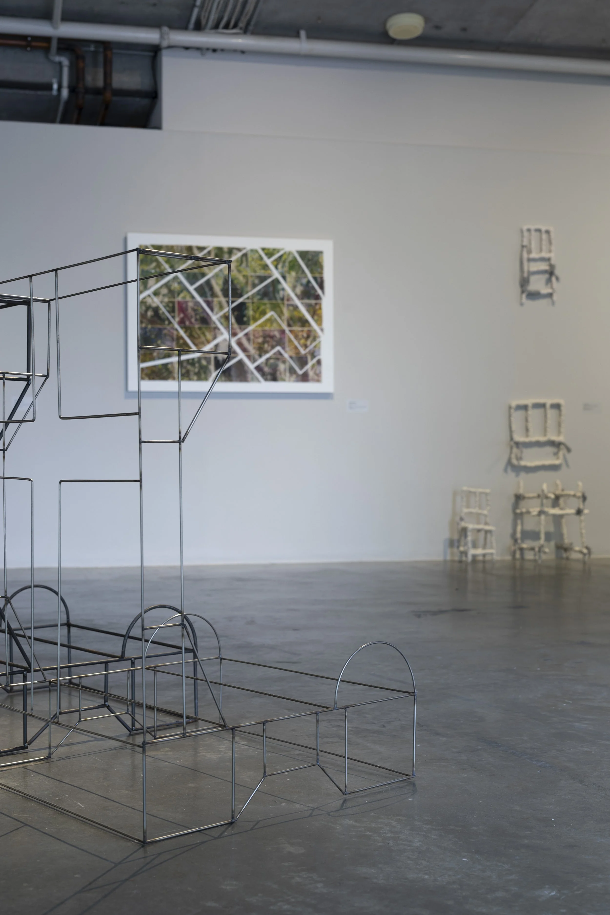 On the floor, metal frame artwork of an alter with a cross on the back. On the wall behind, screenprint of nature with gaps in a geometric pattern and abstract ceramic sculptures akin to chair and crib frames.