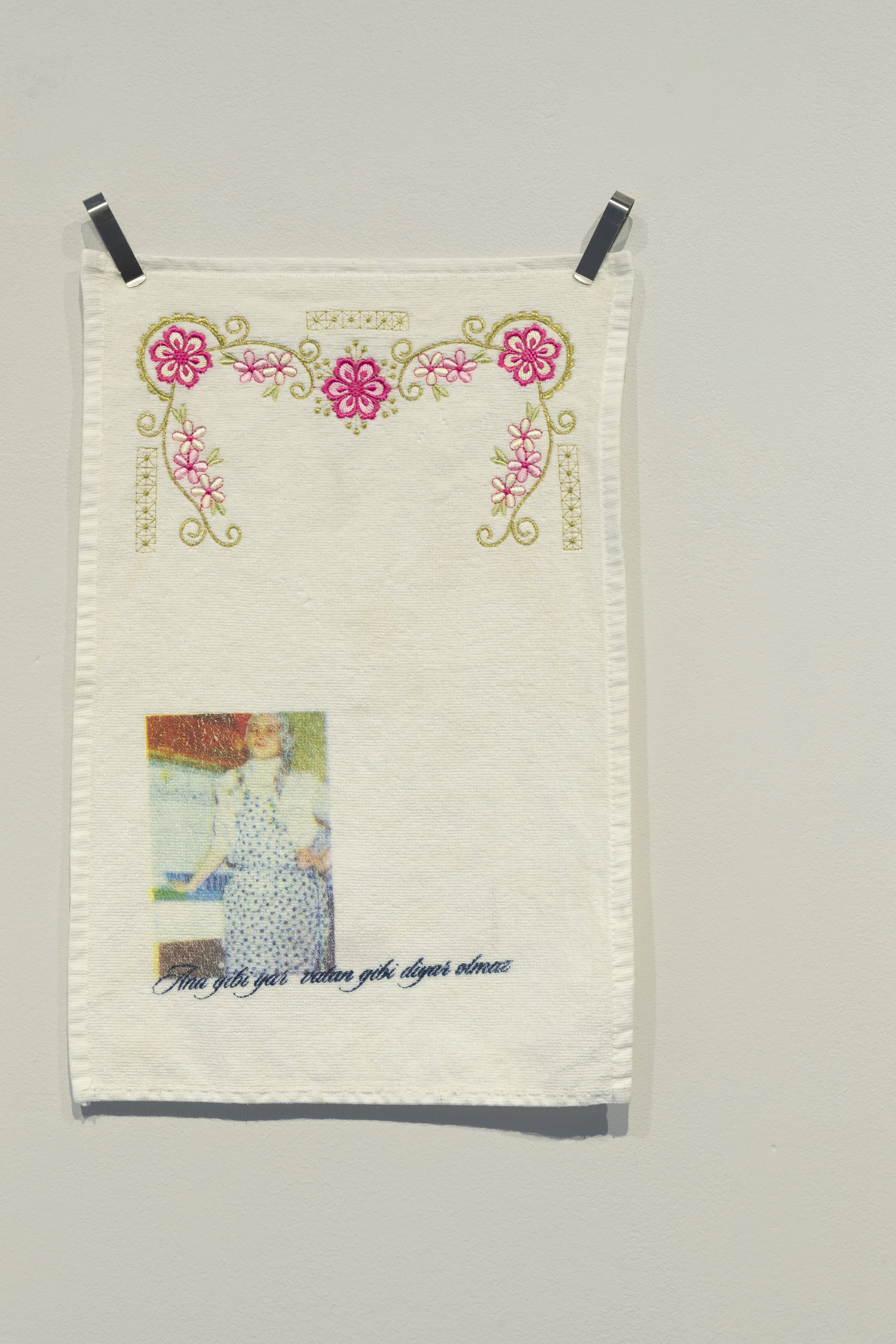 Hanging textile work on wall. Print of a woman standing at a counter and Turkish text in the bottom left corner of a white towel, with pink and green floral embroidery at the top.