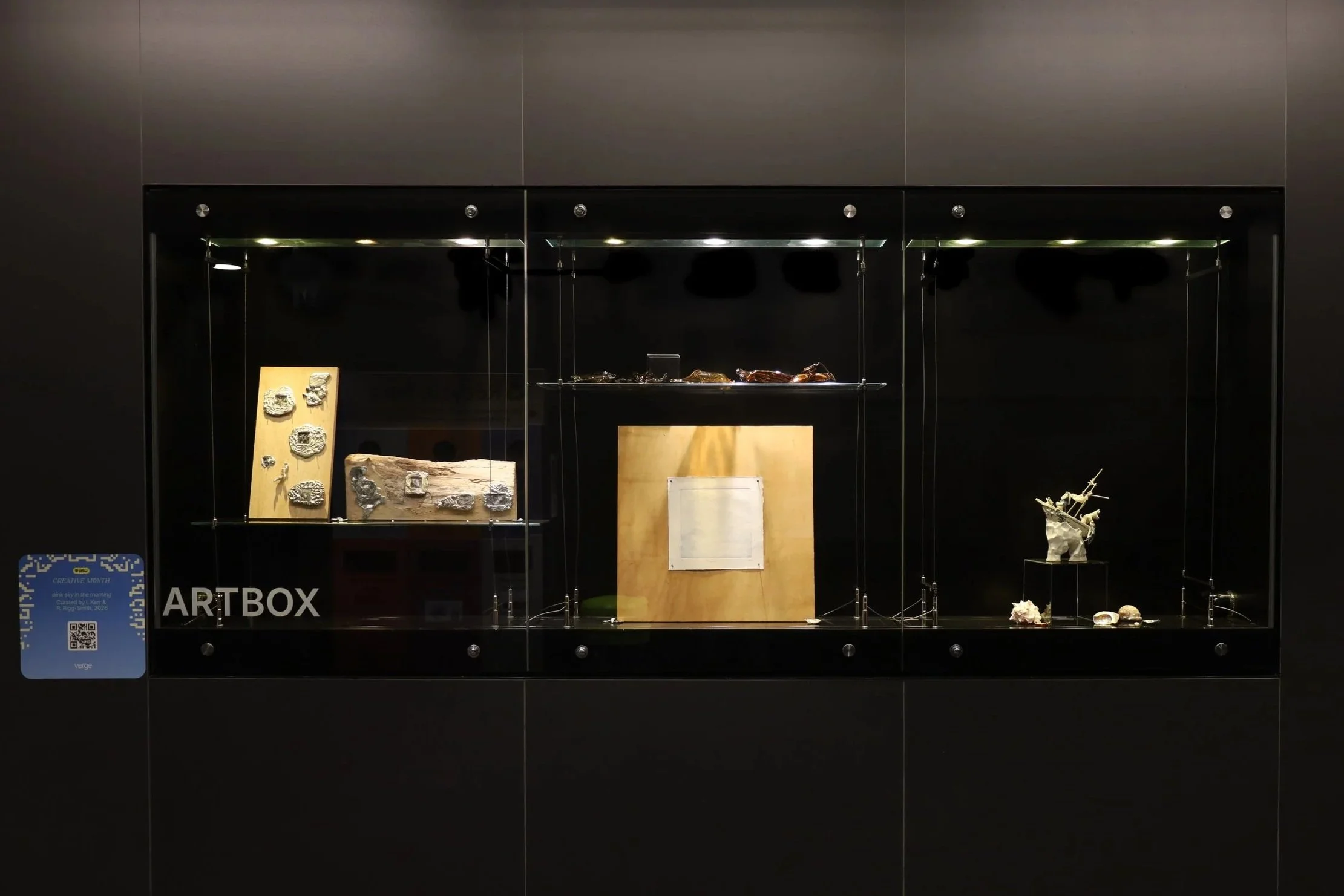 Installation view, left to right. Textured metal sculptures with photographs on wood. Brown blown glass sculptures. Indented paper on square wood board. Small ceramic sculpture of a sinking ship. Shells.