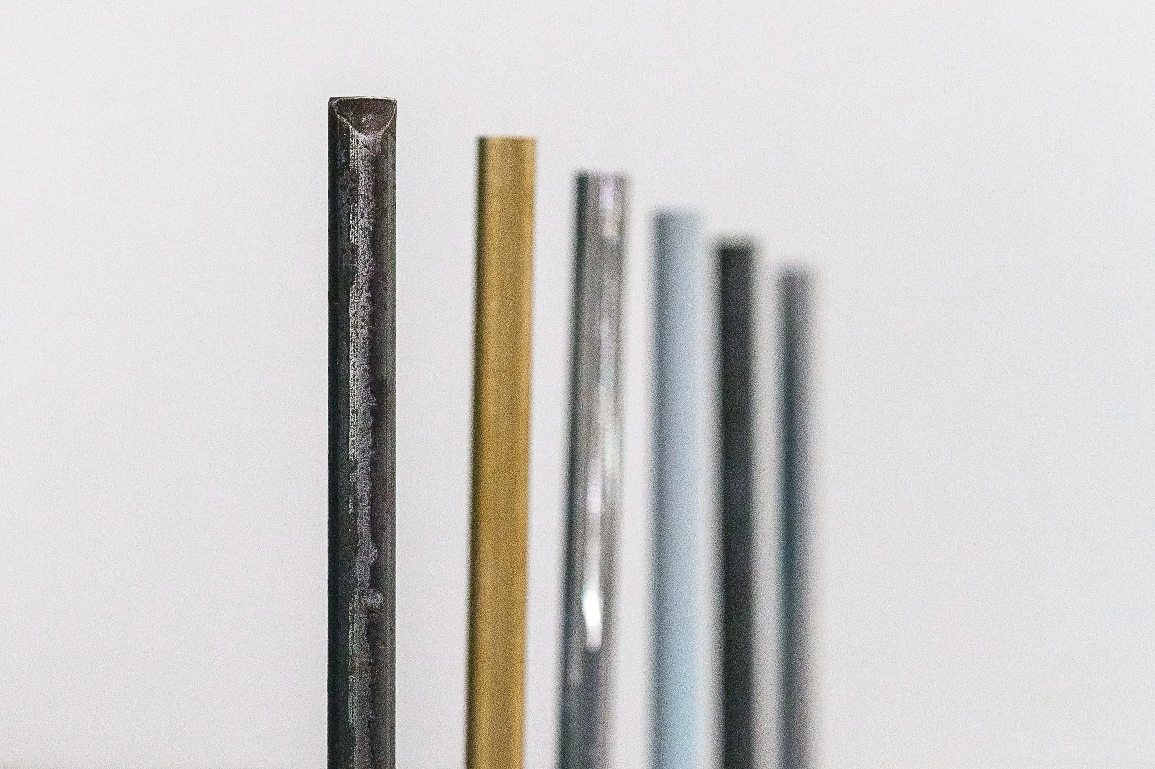 Close up shot of 6 rod like objects in a metal-like material standing in a row. Colours of black, gold, silver, blue. The image becomes blurry toward the back.