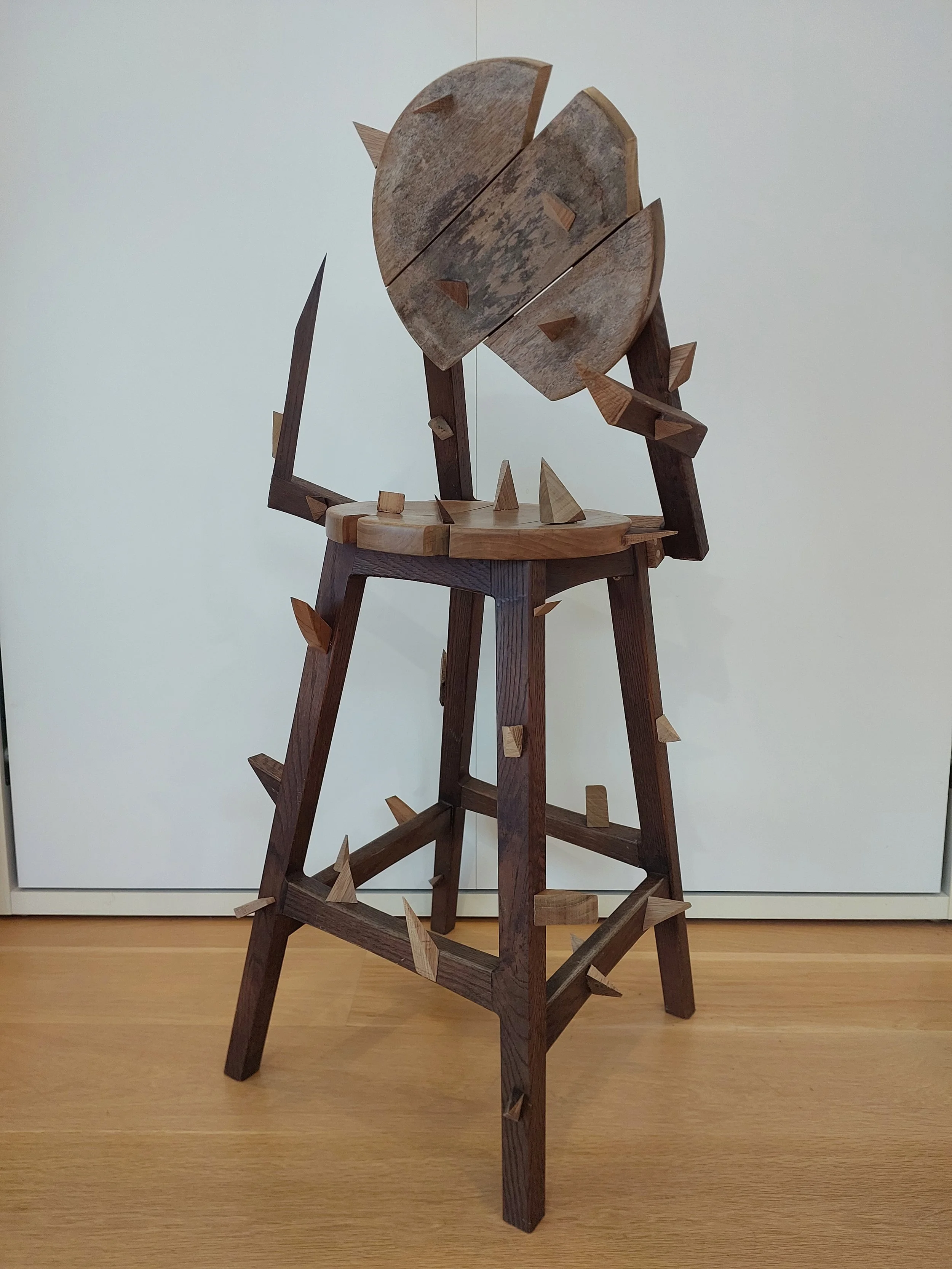 Cian Stroemer, The Chair, 2026. Wooden sculpture of a distorted bar stool. Sharp triangles obtrude from the chair like thorns, and the chair back and seat are segmented and bent.
