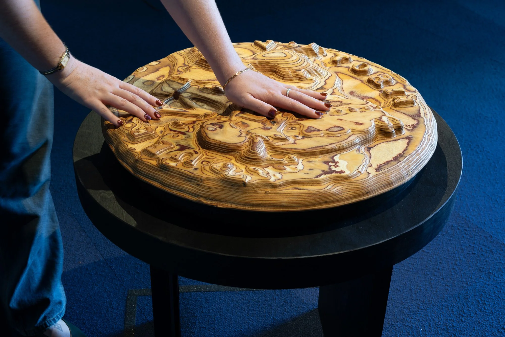 Image: Cindy Yuen-Zhe Chen, Touching Places Past and Present - Penang, 2026. Hands touching circular plywood sculpture on a black plinth. Carved grooves with ridges that resemble text on a coin as well as land topography.