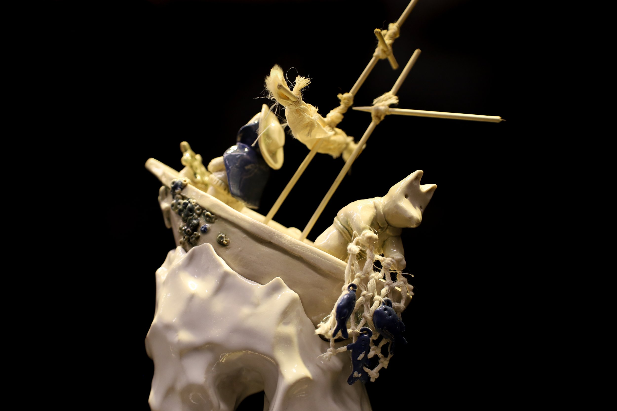 Ceramic sculpture in white, depicting 2 small animal figures on a ship. One wears a hat, steering the ship, the other holding a net full of fish. The ship is on a high wave, with wooden masts and black ceramic barnacles.