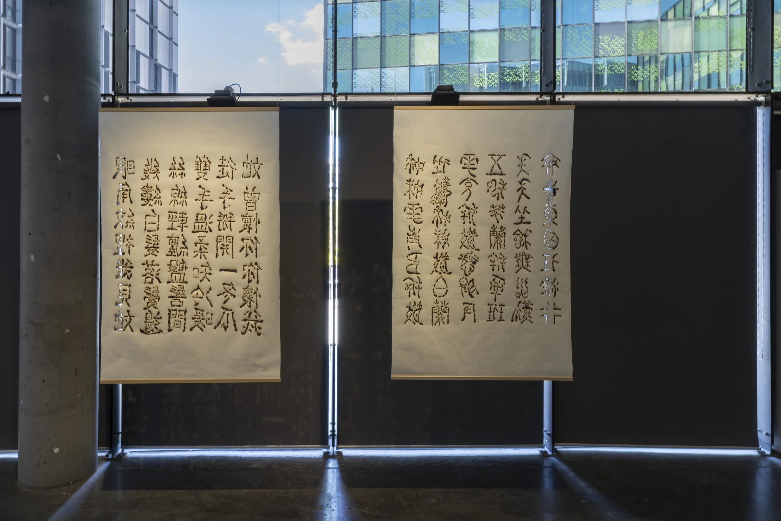 2 Suspended sheets of off-white paper, hung with rods. Each sheet has 6 columns of calligraphy made with perforated burn marks, leaving holes one can see through, and the edges jagged and brown.