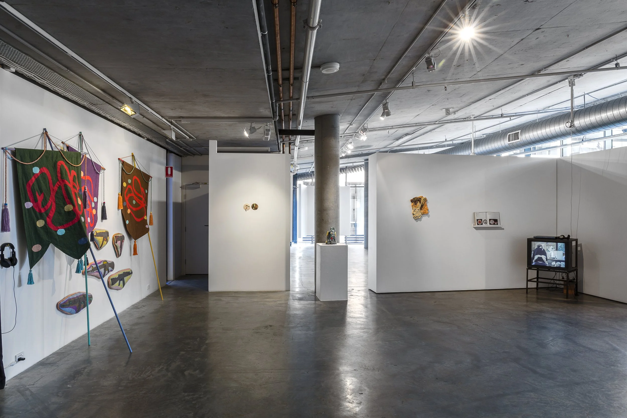 Left to right. Assemblage of banners on sticks with tassels and cushions with guns embroidered. 2 small circular works on the wall. Ceramic sculpture on plinth and wall. Open book on shelf. CRT with video of figure sitting.
