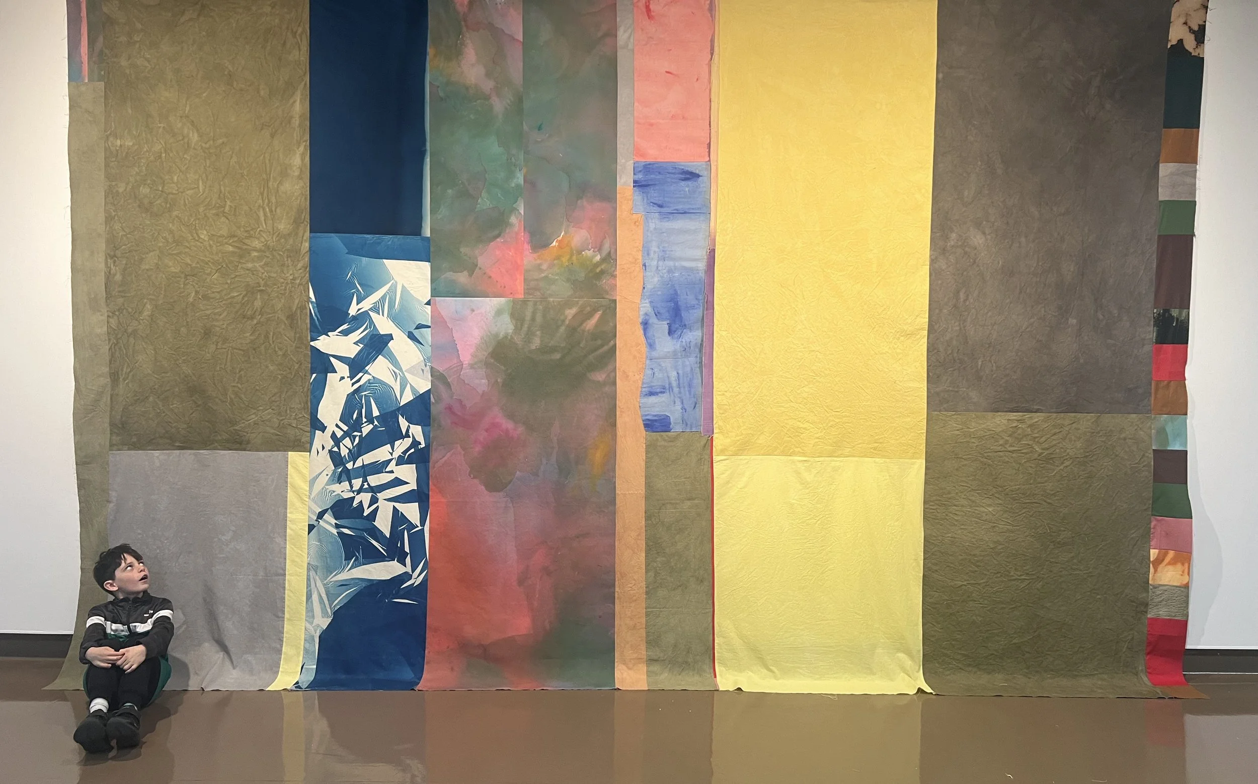 Image: Kylie Banyard, Soft wall 2, 2024. Large hanging tapestry with patch work fabrics in blocks and vertical stripes. Colours of yellow, dark green, blue and pink. Some patterned, others plain. Artist's son sits in corner.