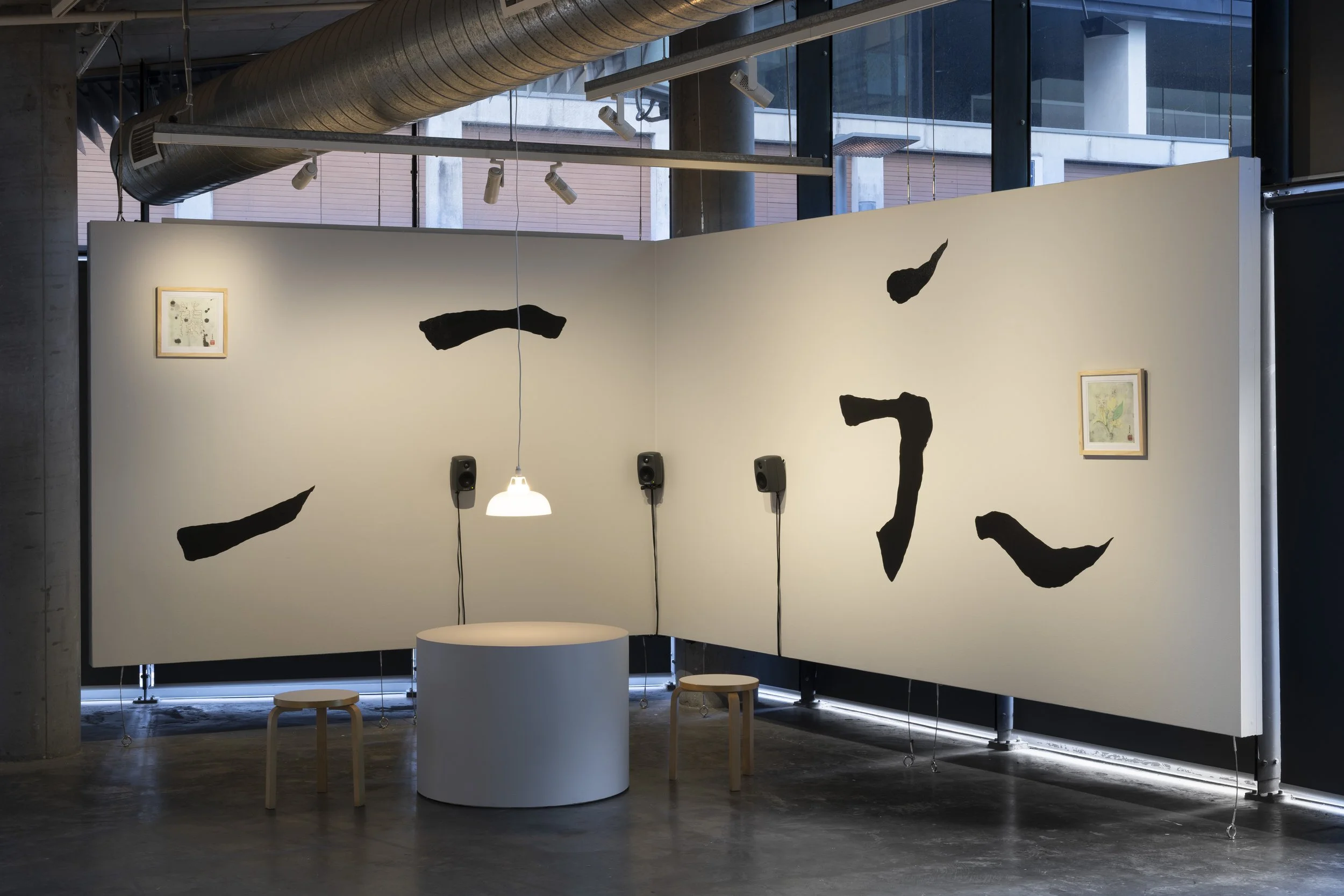 Corner of gallery with mural of fragmented black strokes on white wall. 2 square, framed watercolour paintings of orchids on either end. Center, white cylinder table, 2 stools and lamp hanging above, 3 small speakers behind.