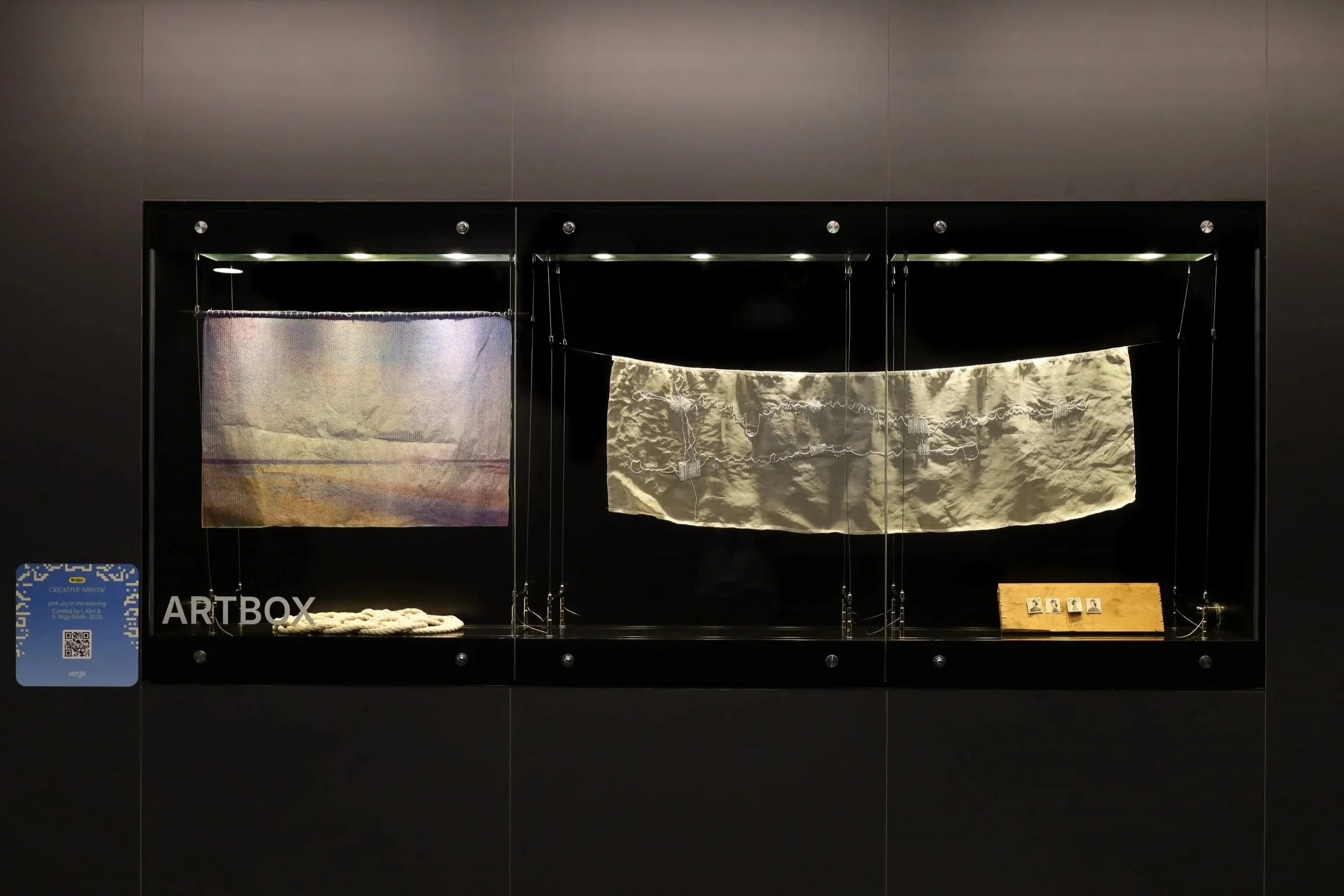 Installation view, left to right. Hanging pastel screen print on a wooden stick. Patterned knot of thick rope. Hanging white embroidered tapestry. 4 small black and white photographs nailed onto a wood plank.