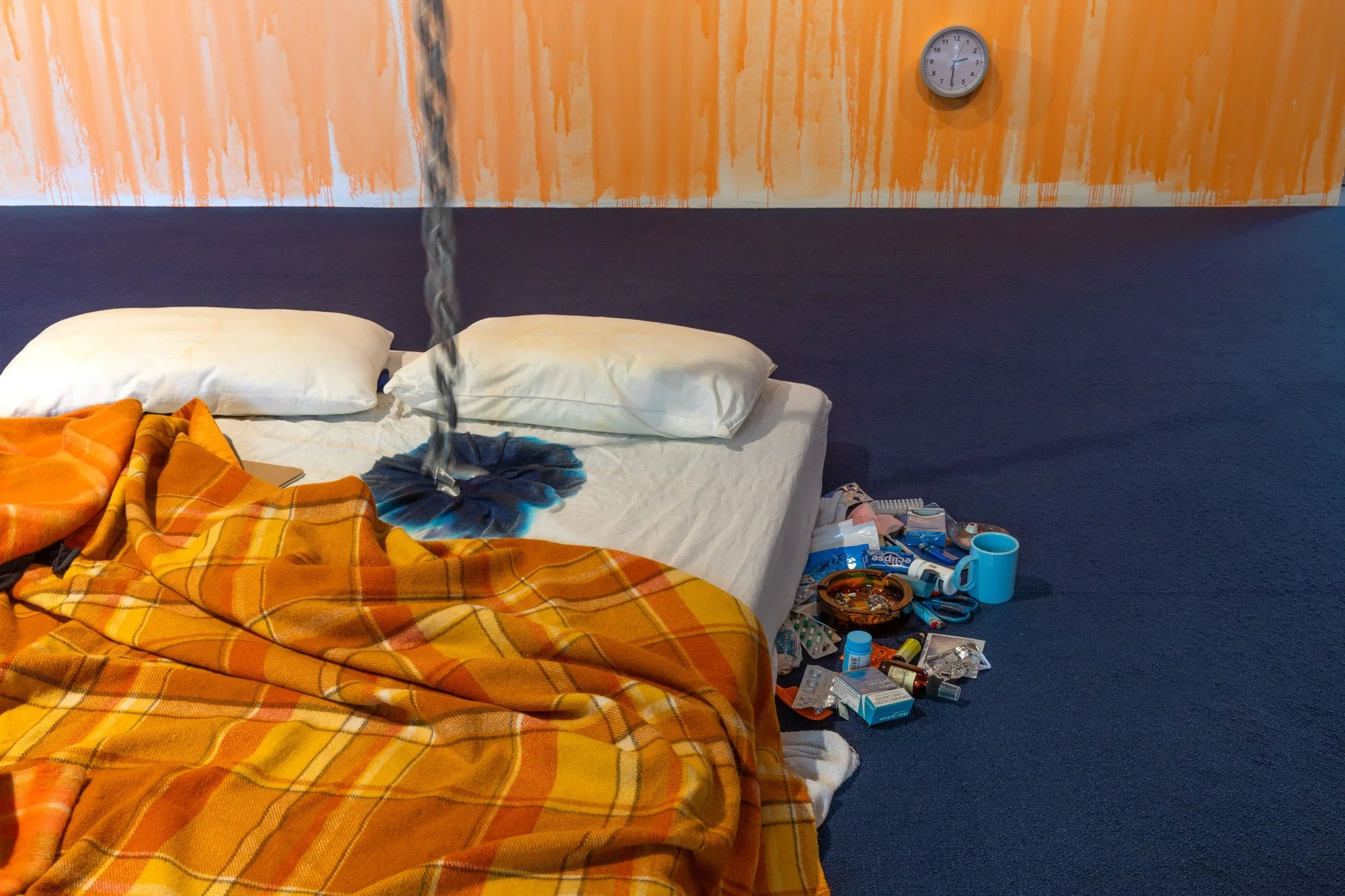 Installation view of Sixty-nine by Sidney McMahon, 2025. Mattress with orange bedding on navy carpet with personal items on the floor. Chain suspended from the ceiling and connected to the bed.