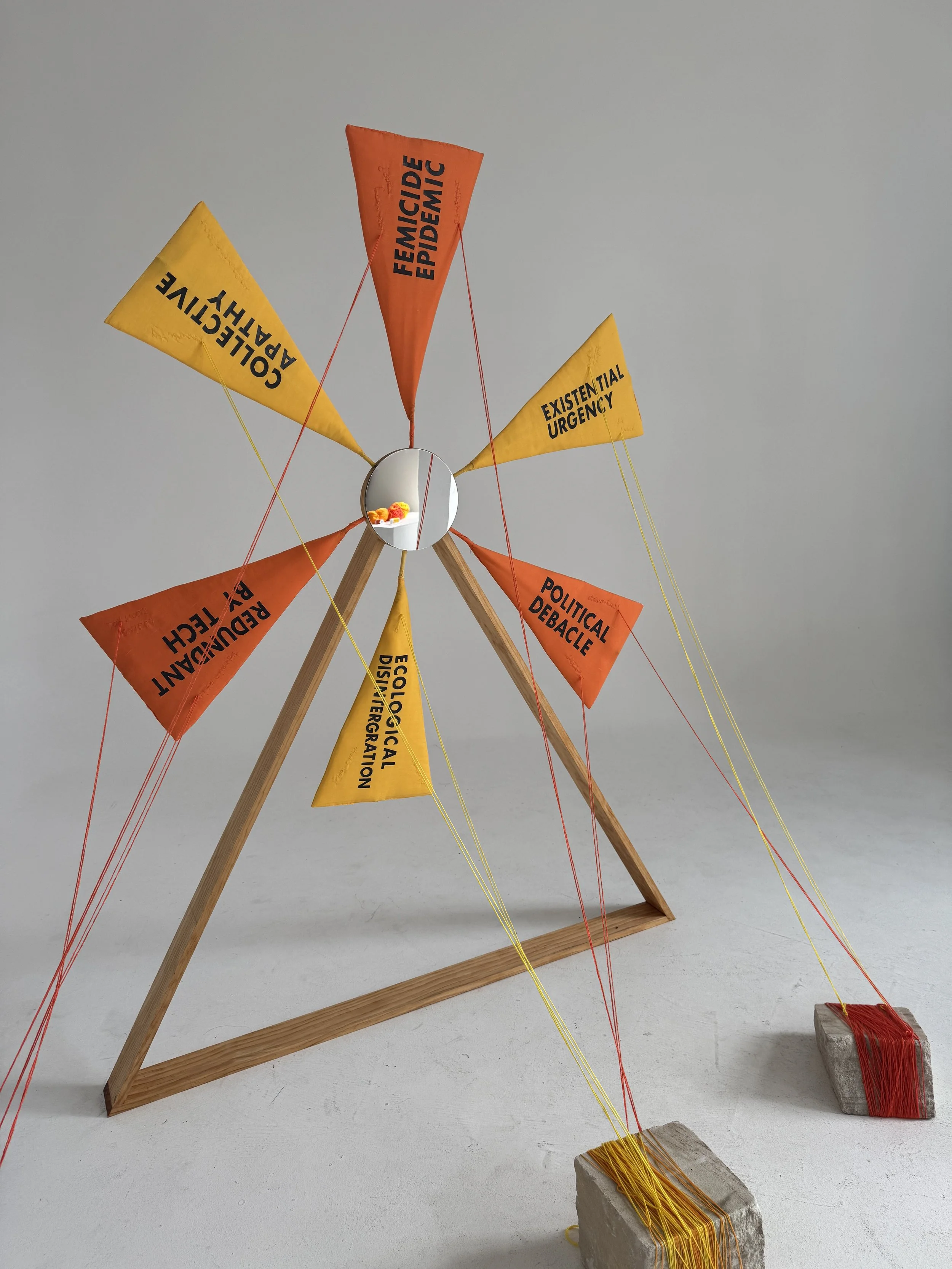 Ruby Stephens-Naughton, Tomorrow's Problem, 2026. Sculpture of an abstracted windmill of wooden frame, mirror and 6 yellow and orange sails with black text. The sails are tied down to sandstone blocks with coloured string.