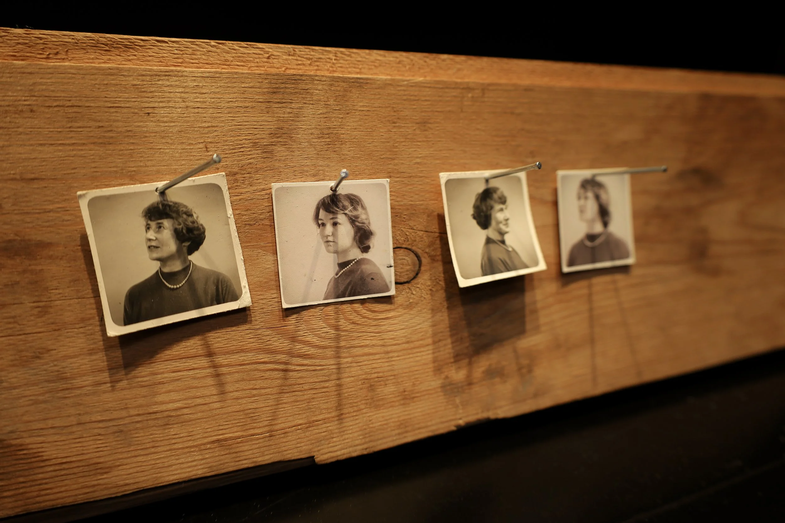 4 small Black and white portraits nailed to a plank of wood. The portraits depict a woman with short hair, wearing a pearl necklace and mock-neck sweater. The photos alternate between an older woman and a young woman.