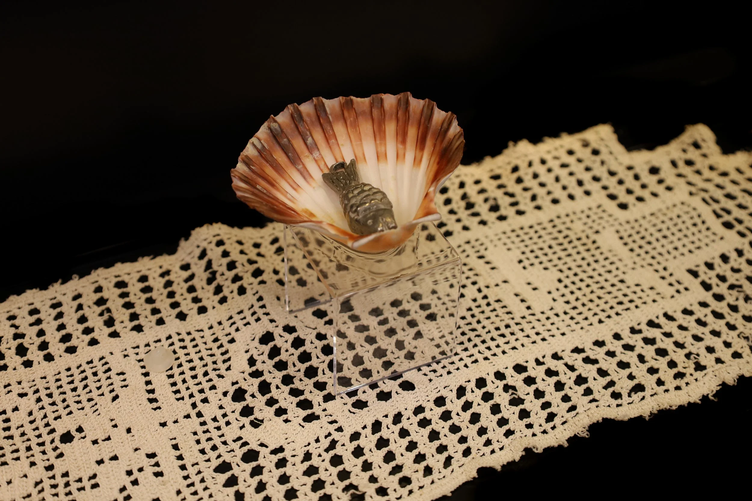 Small pewter fish sculpture in a red and white scallop shell. The shell sits on a clear acrylic stand, on top of a white crocheted mat.