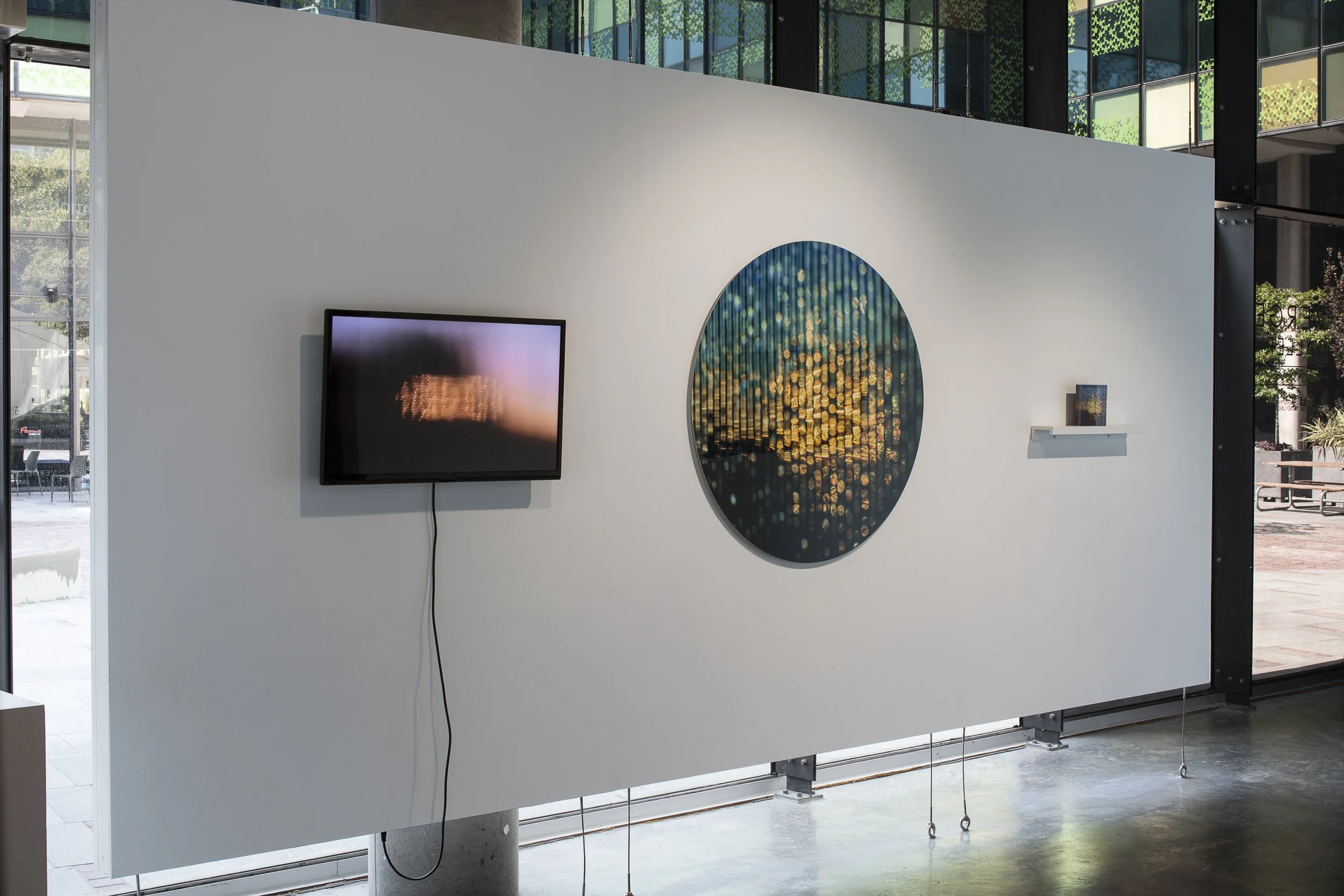 Left, video monitor displaying blurred video in purple, orange and black. Middle, large circular photo of blurred blue, yellow and black dots. Right, Small acrylic block printed with the same image on a shelf.
