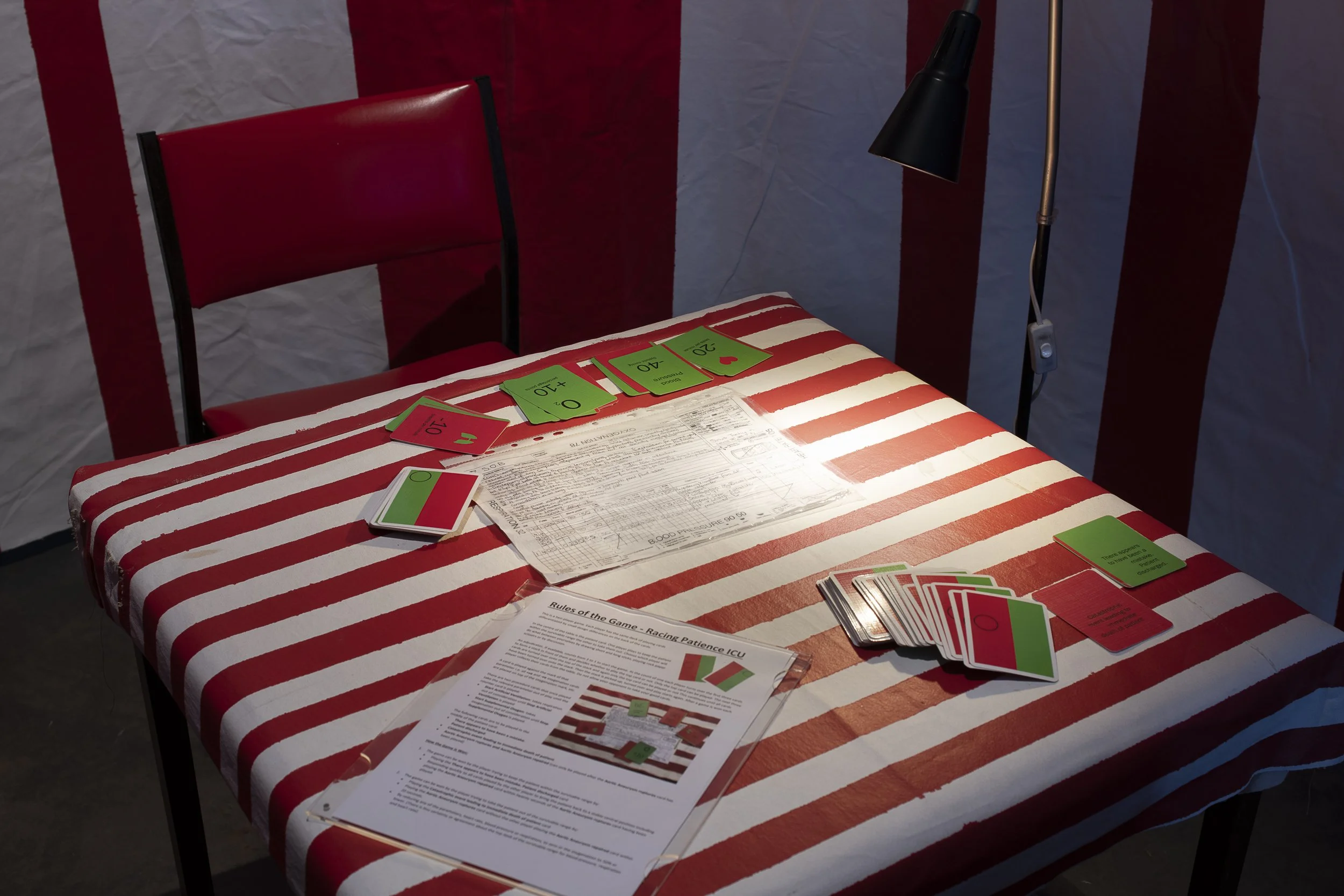 Close up of an installation work. A room space with red and white striped table, chairs and wall. A green and red pack of cards alone with game instructions and medical form are on the table. A lamp beside shines down on the tabletop.