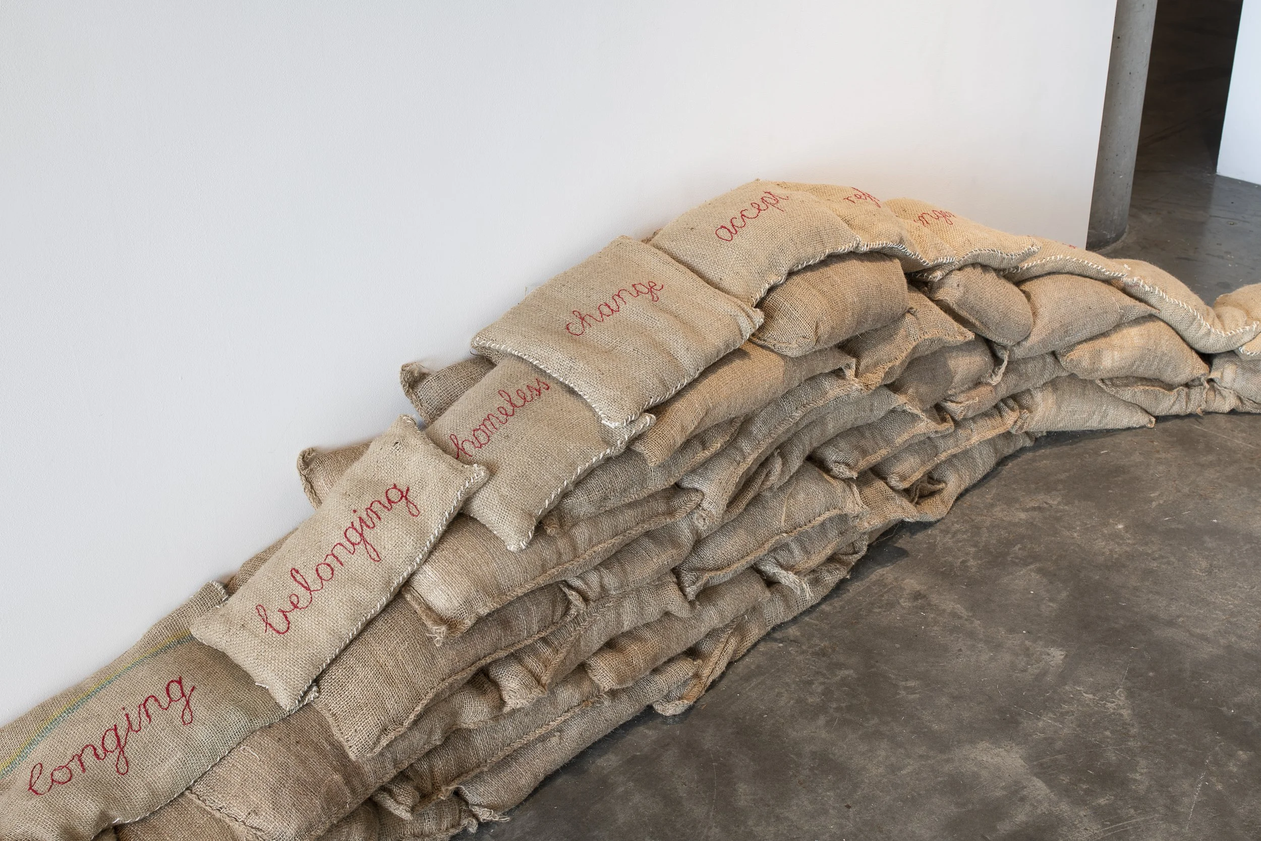 Close up on the sandbags - the ones at the top have red embroidered cursive words, some reading: "longing, belonging, homeless, change, accept...".