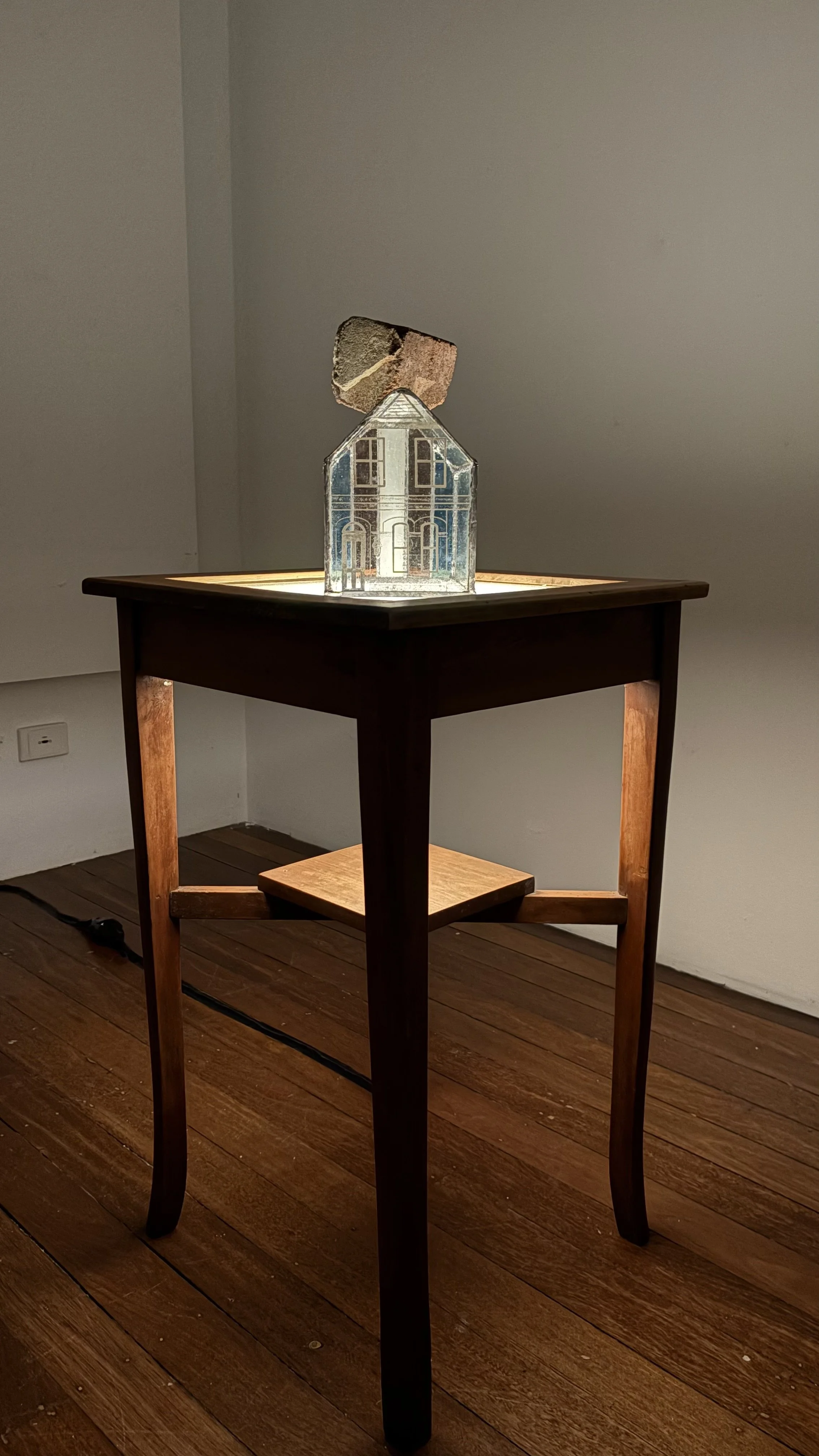 Xavier Marz, UNDER PRESSURE, 2026. Sculpture of a glass house with a large brick on its roof, illuminated by a light below. The sculpture sits on a tall wooden table.