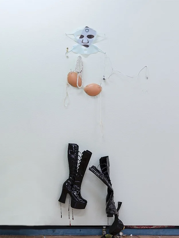 Image: Bonnie Huang, jesus crucified on the cross, 2022. Found object assemblage of face mask, silicone breast cups and boots nailed to the wall.