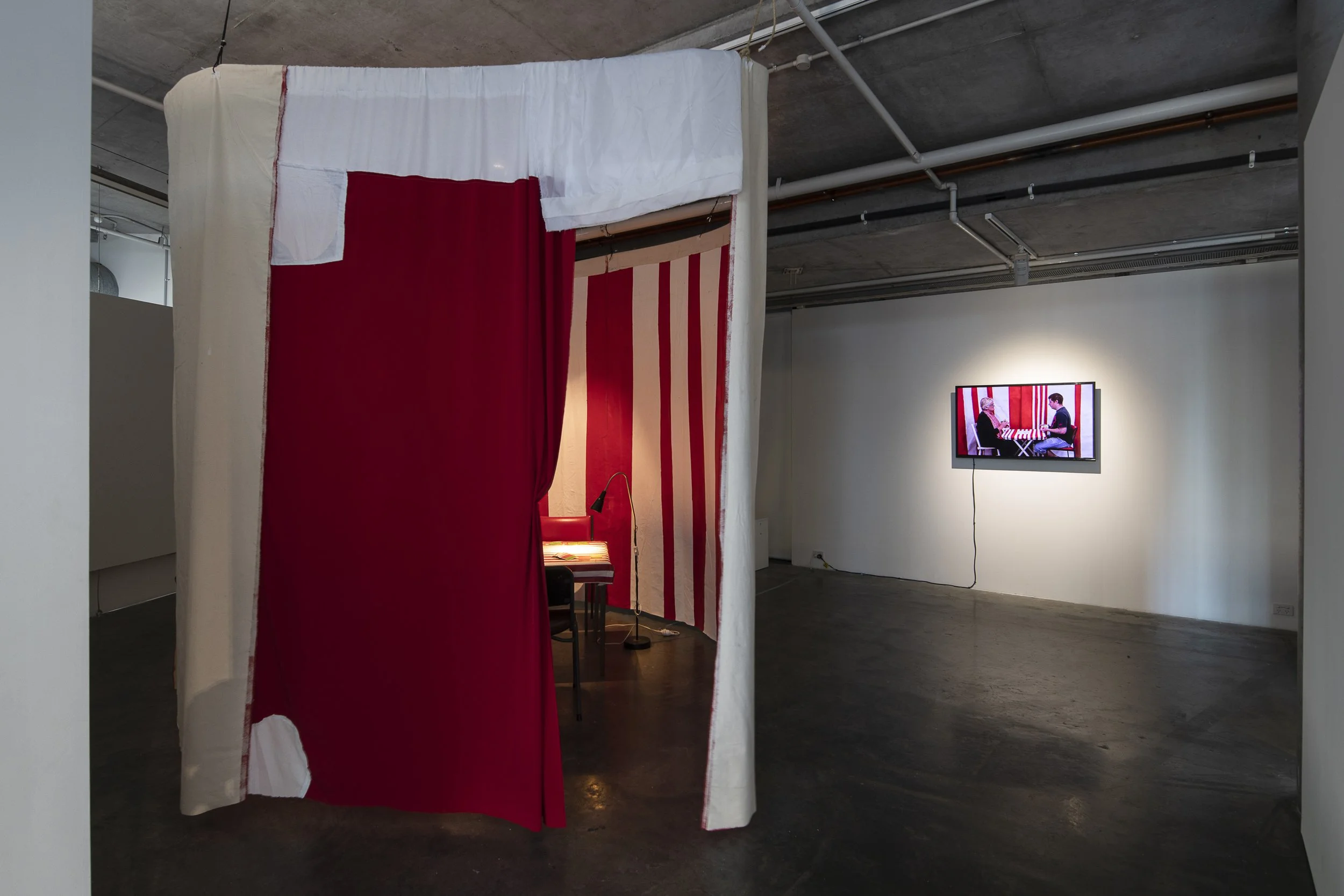 Left, Red and white striped fabric is suspended to create a room like space with a table, chairs and a lamp inside. Right, video monitor displaying two people sitting in a red and white striped room.