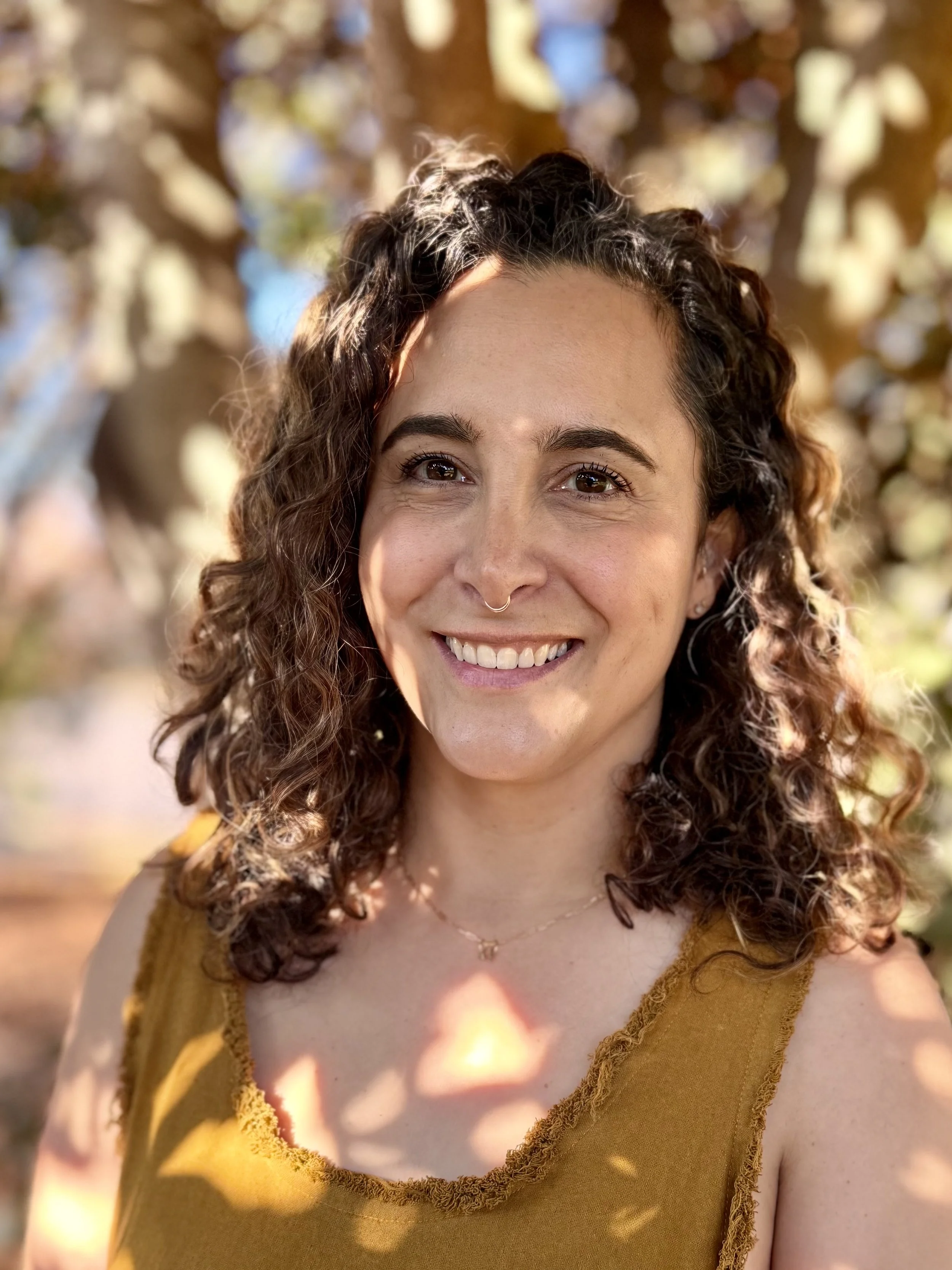 A woman with curly brown hair smiling outdoors in autumn, wearing a mustard yellow sleeveless top with a textured trim around the neckline and a thin gold necklace.