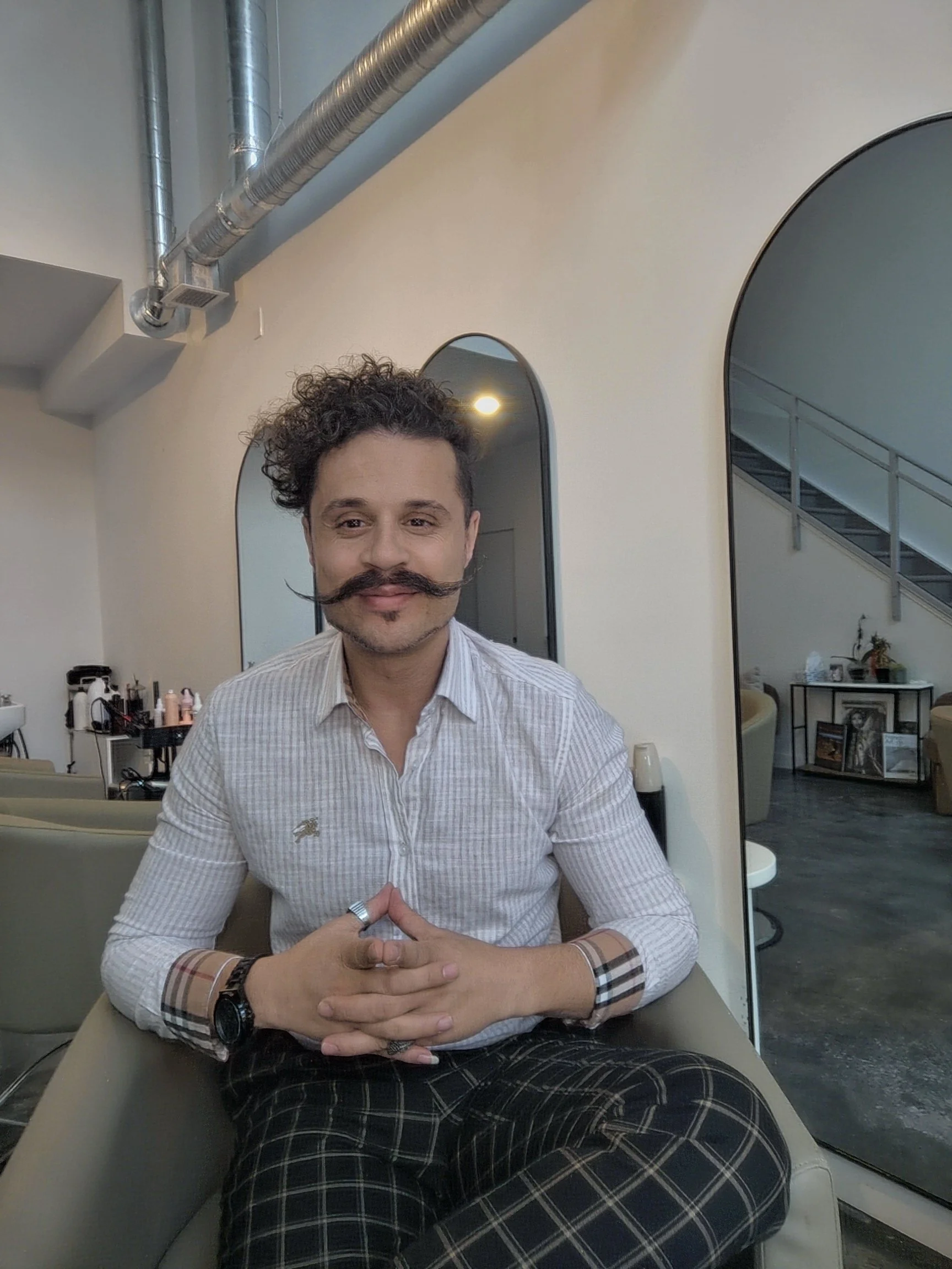 A man with curly hair and a styled mustache is sitting on a chair with his hands clasped. He is wearing a light-colored, checked shirt and plaid pants. The background shows a modern interior with mirrors, stairs, and some furniture.