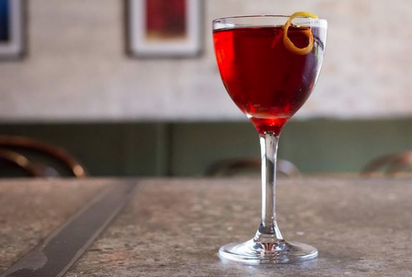 A glass of red cocktail with a lemon twist garnish on a bar counter