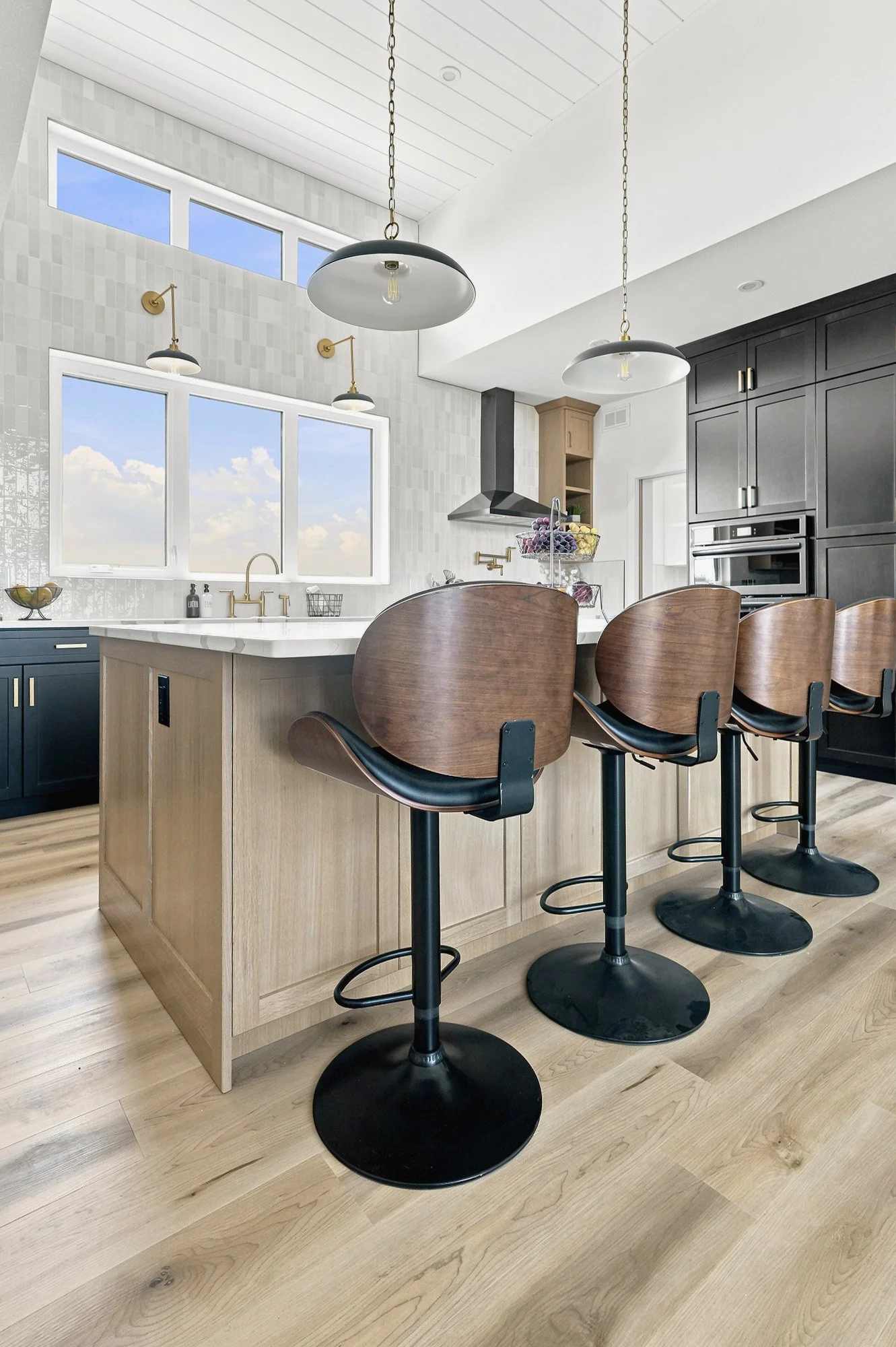 Modern kitchen with wood island, black chairs, pendant lights, large windows, and light wood flooring.
