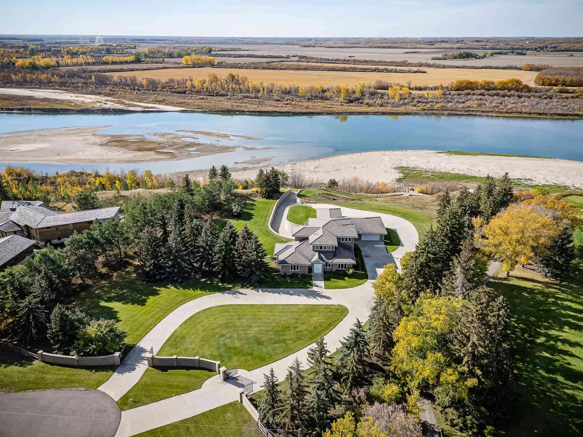 A real estate aerial view of a large house with a circular driveway, surrounded by trees, near a river and scenic fields.