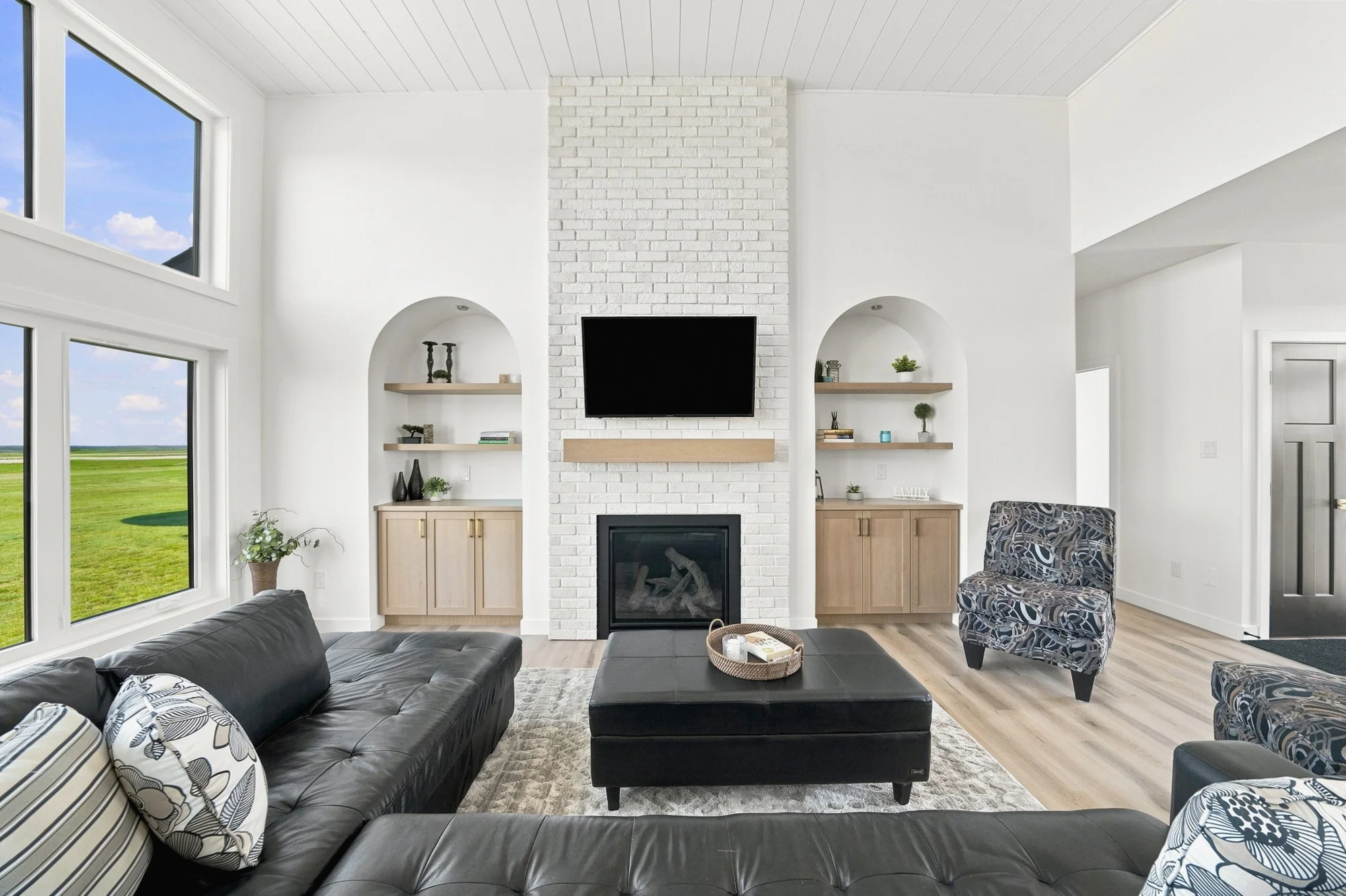 Modern living room with black furniture, a white brick fireplace, wall-mounted TV, built-in shelves, and large windows.