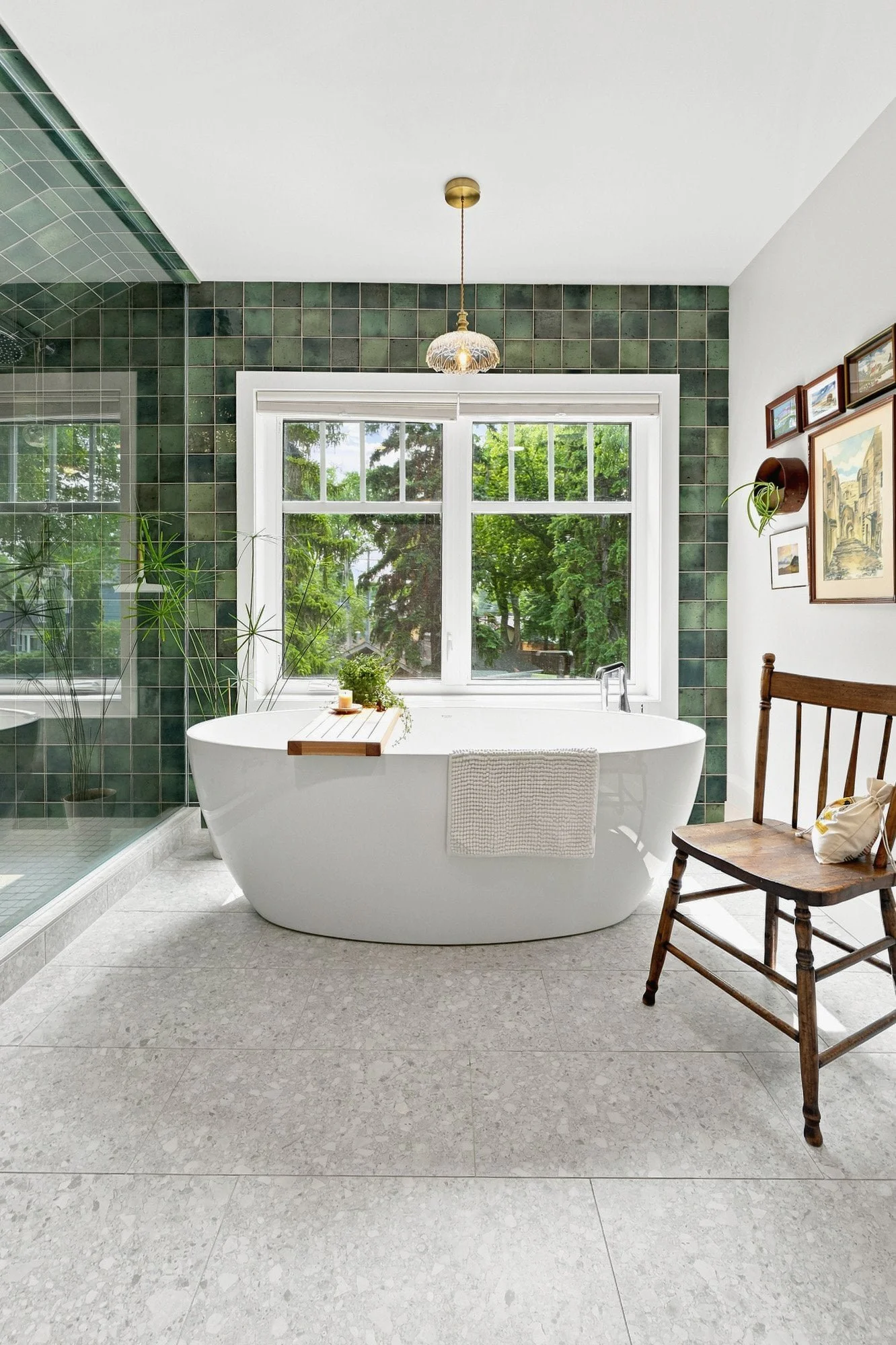 Modern bathroom with a white tub, green tile wall, large window, wooden chair, and potted plants.