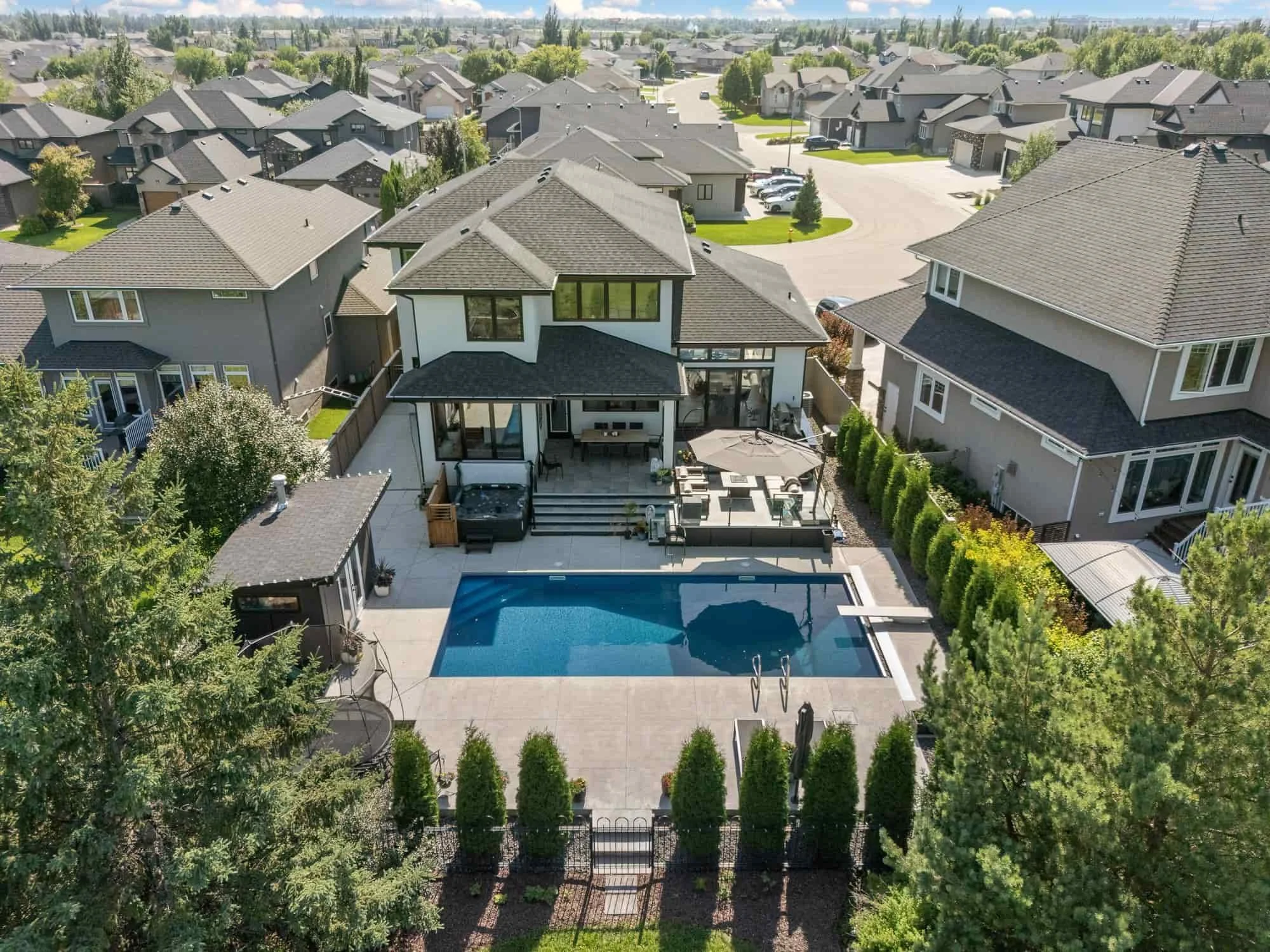 Aerial real estate drone photography captures a modern house with a pool, patio furniture, and lush landscaping in a suburban neighborhood.