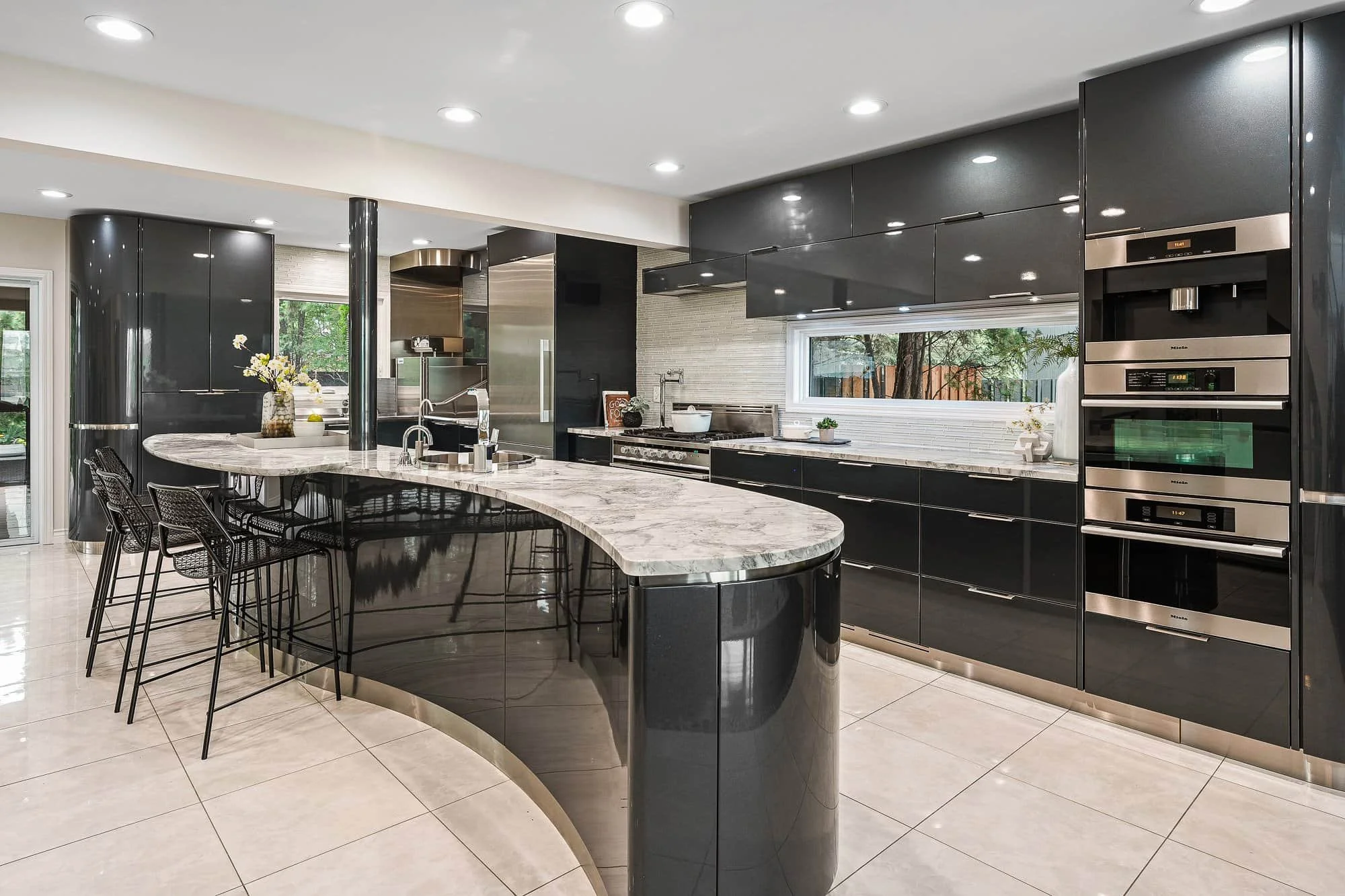 Modern kitchen with glossy black cabinets, marble curved island, built-in ovens, and a large window—perfect for real estate photography.
