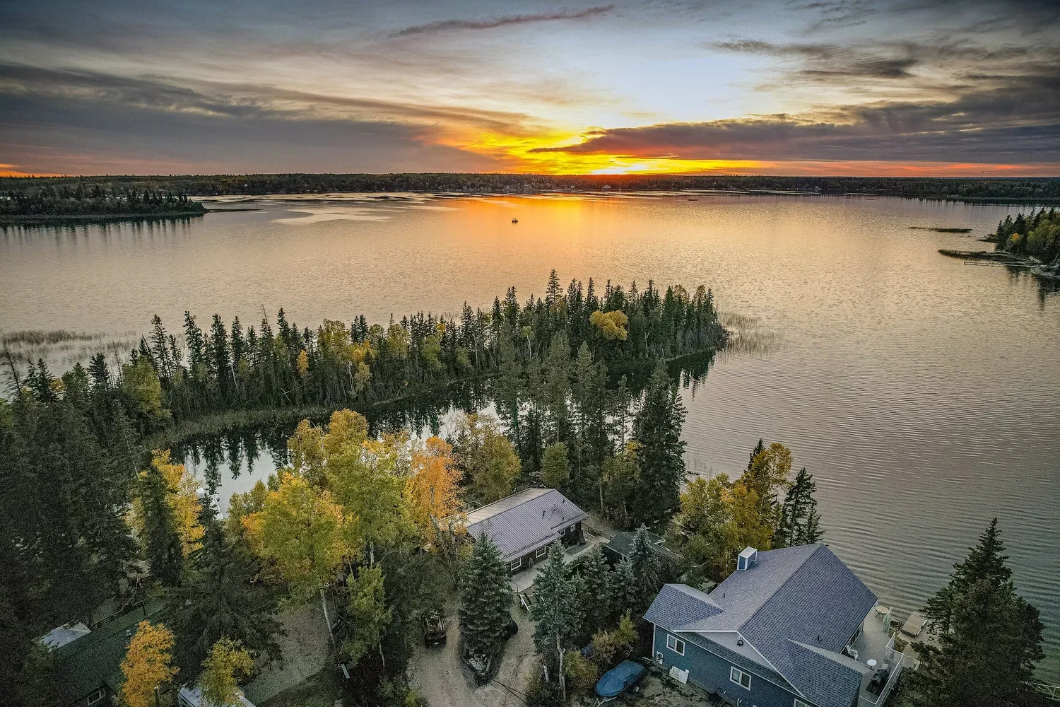 Drone view of a lakeside cabin at sunset surrounded by trees and calm water.