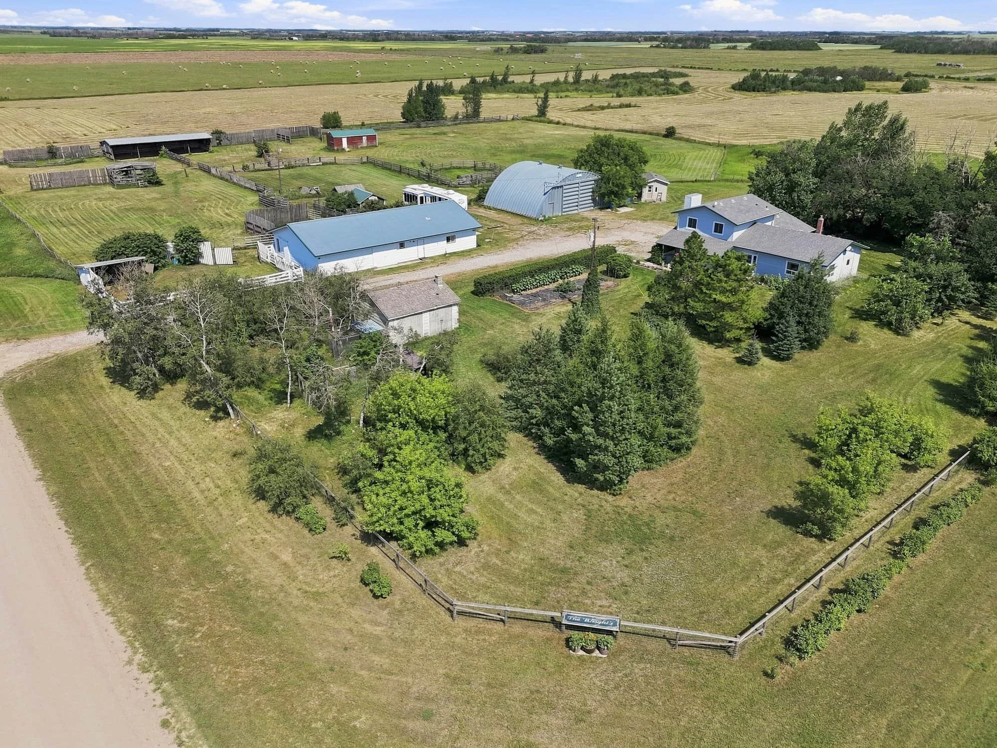 A rural farm with barns, sheds, fields, trees, and a house surrounded by grass and a wooden fence, viewed from above.