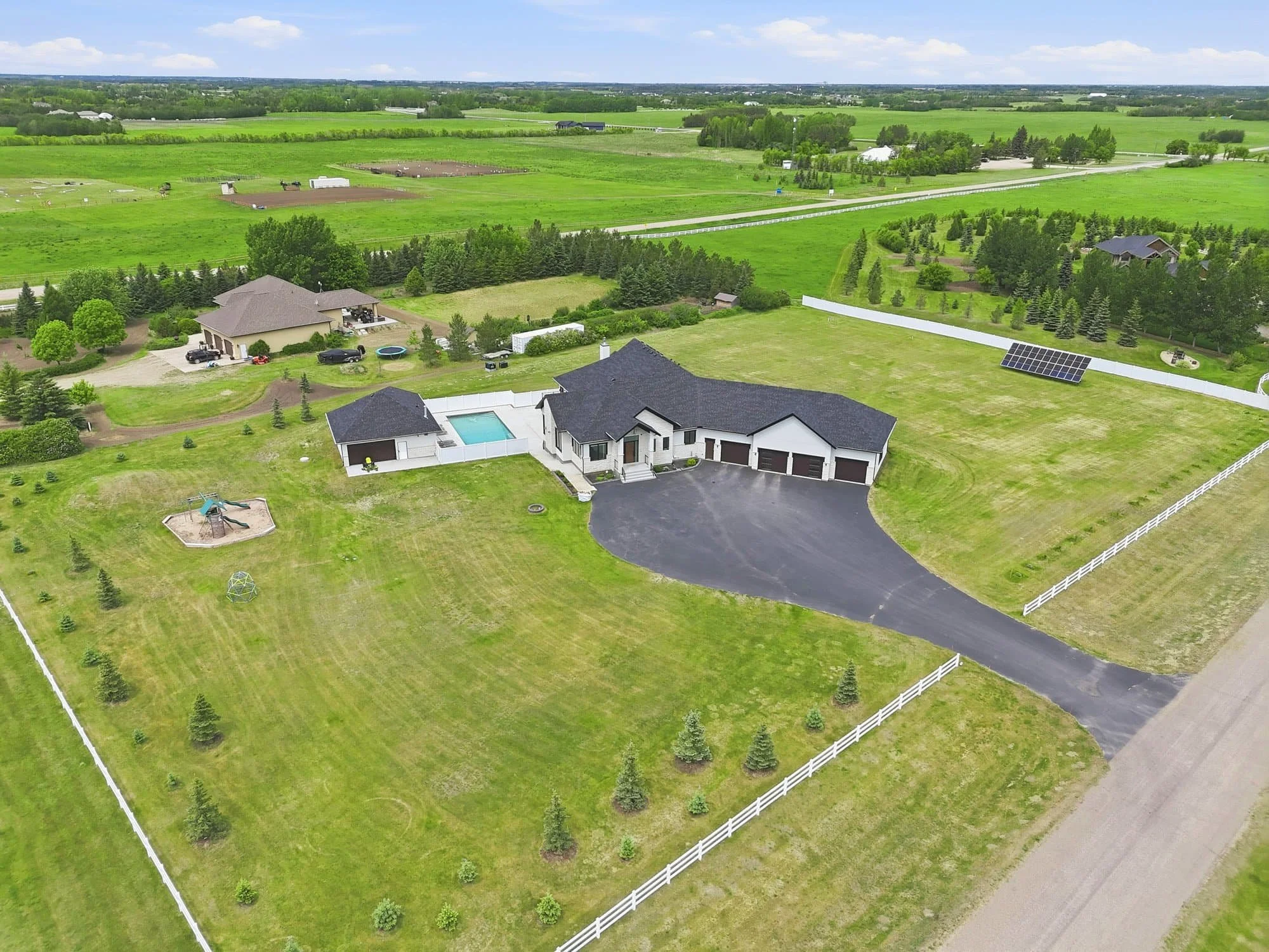Aerial view of a large house with a pool, driveway, and fenced yard surrounded by green fields and trees.