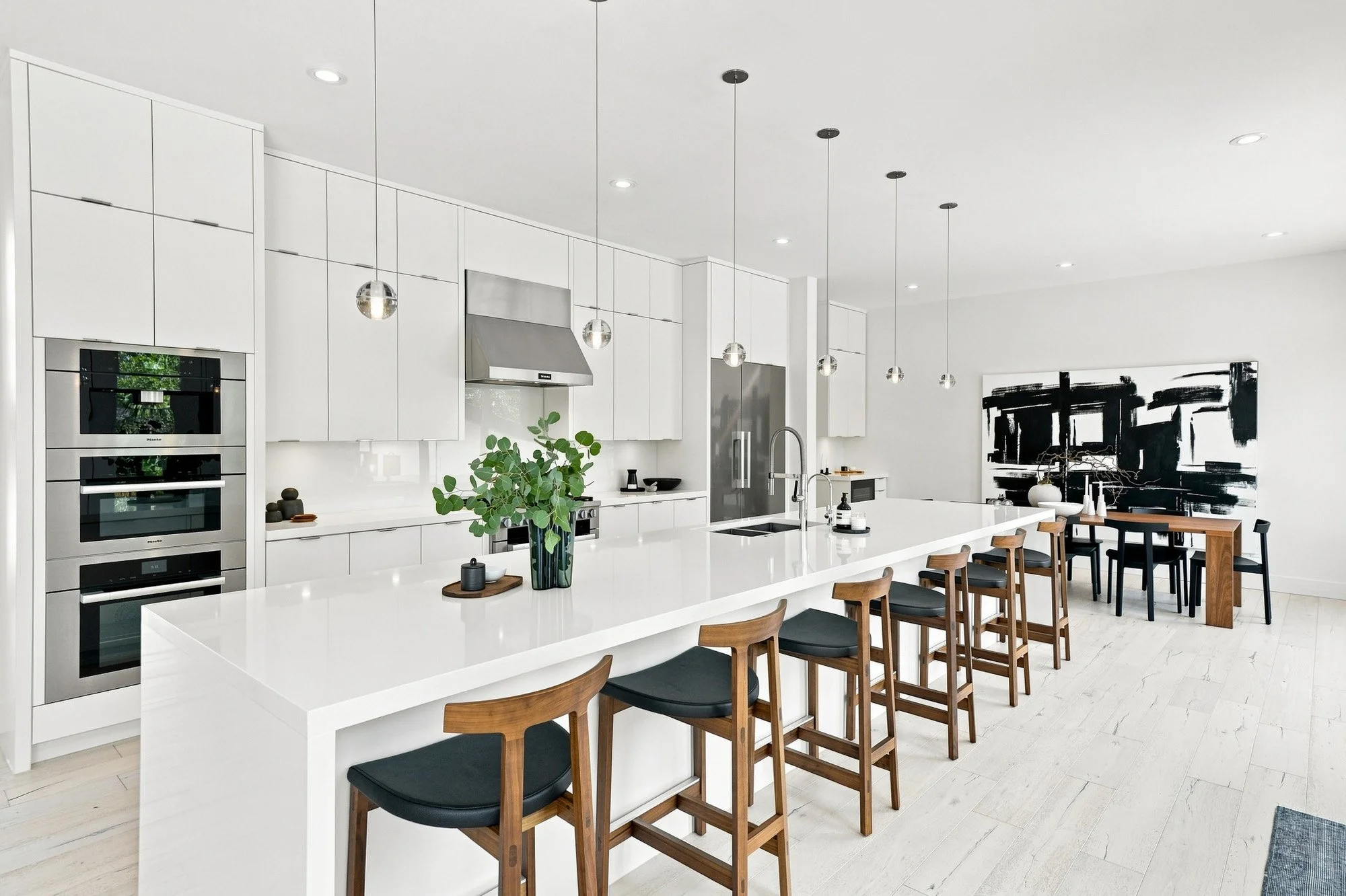 Modern white kitchen with long island, wooden stools, pendant lights, and abstract art on the far wall.