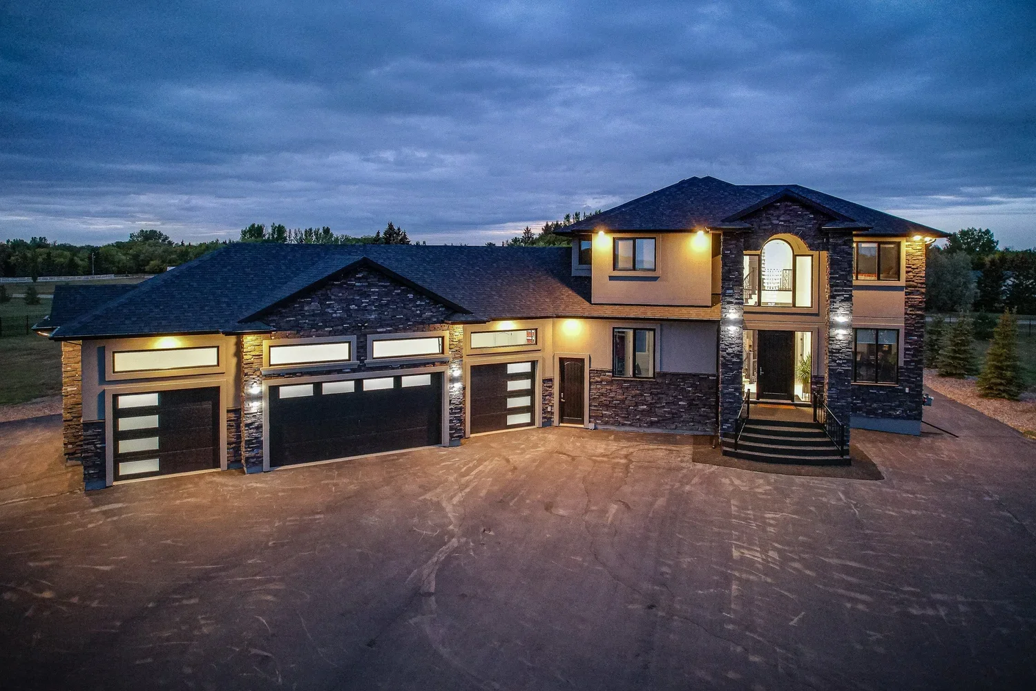 Drone view of a luxury home's front exterior at dusk with warm exterior lighting.