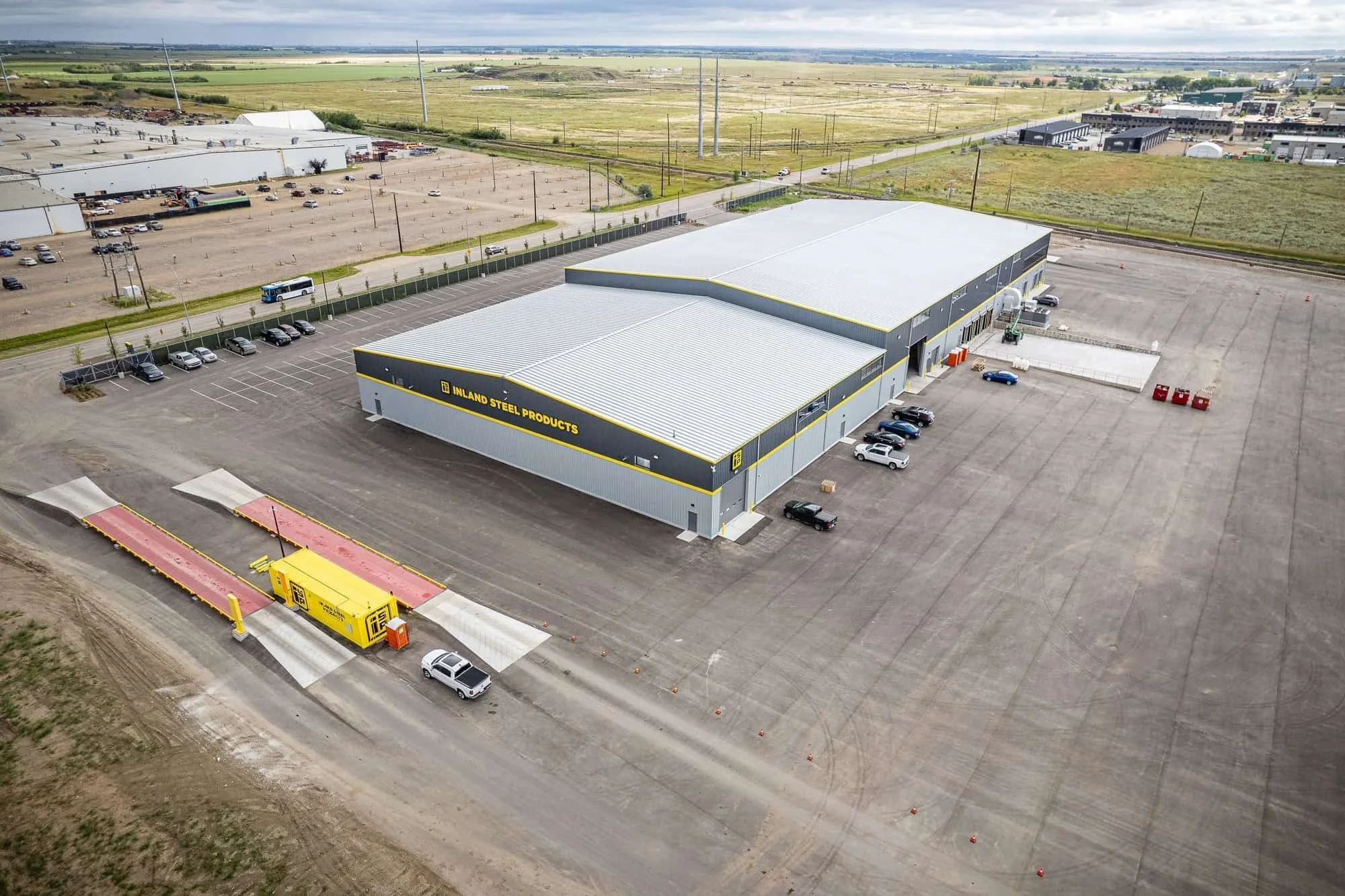 A large industrial warehouse with cars and trucks parked outside, perfect for industrial photography amid open fields and a paved lot.
