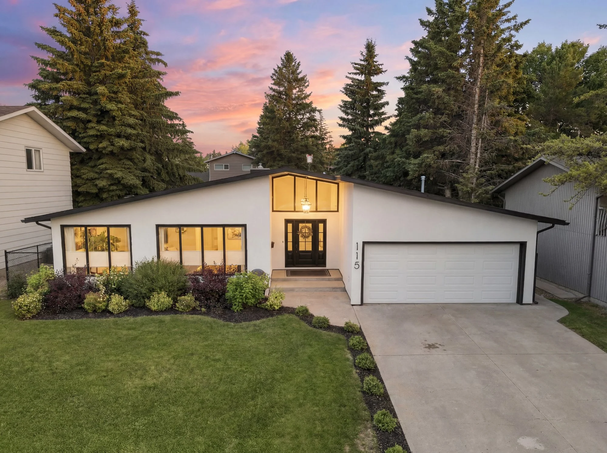 Twilight exterior of a modern Saskatoon home with illuminated windows, mature trees and landscaped front yard