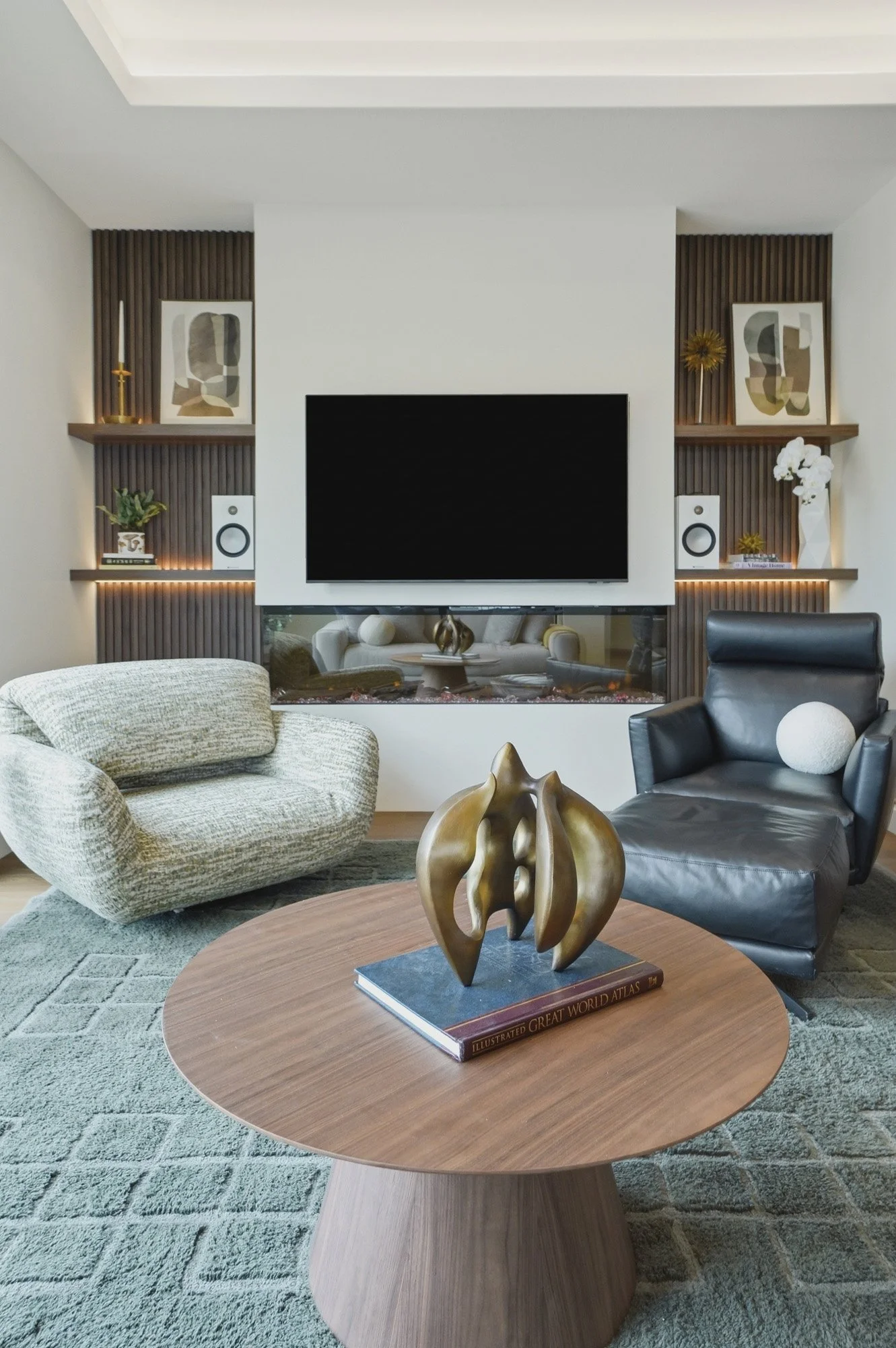 Modern living room with cozy chairs, a round table with abstract sculpture, TV, art, speakers, and shelf decor.