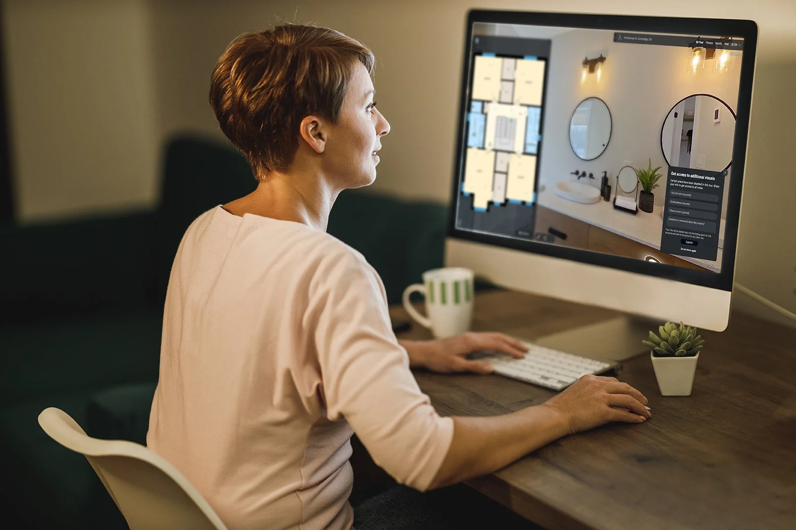 A woman sits at a desk using a computer to view virtual tours of homes, with a mug and small plant beside her.