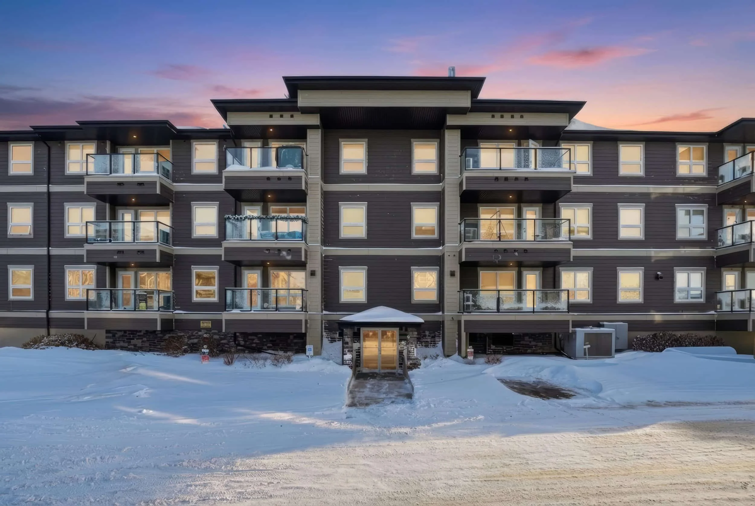 A modern three-story apartment with balconies glows in virtual twilight photography, snowy ground shimmering at sunset.