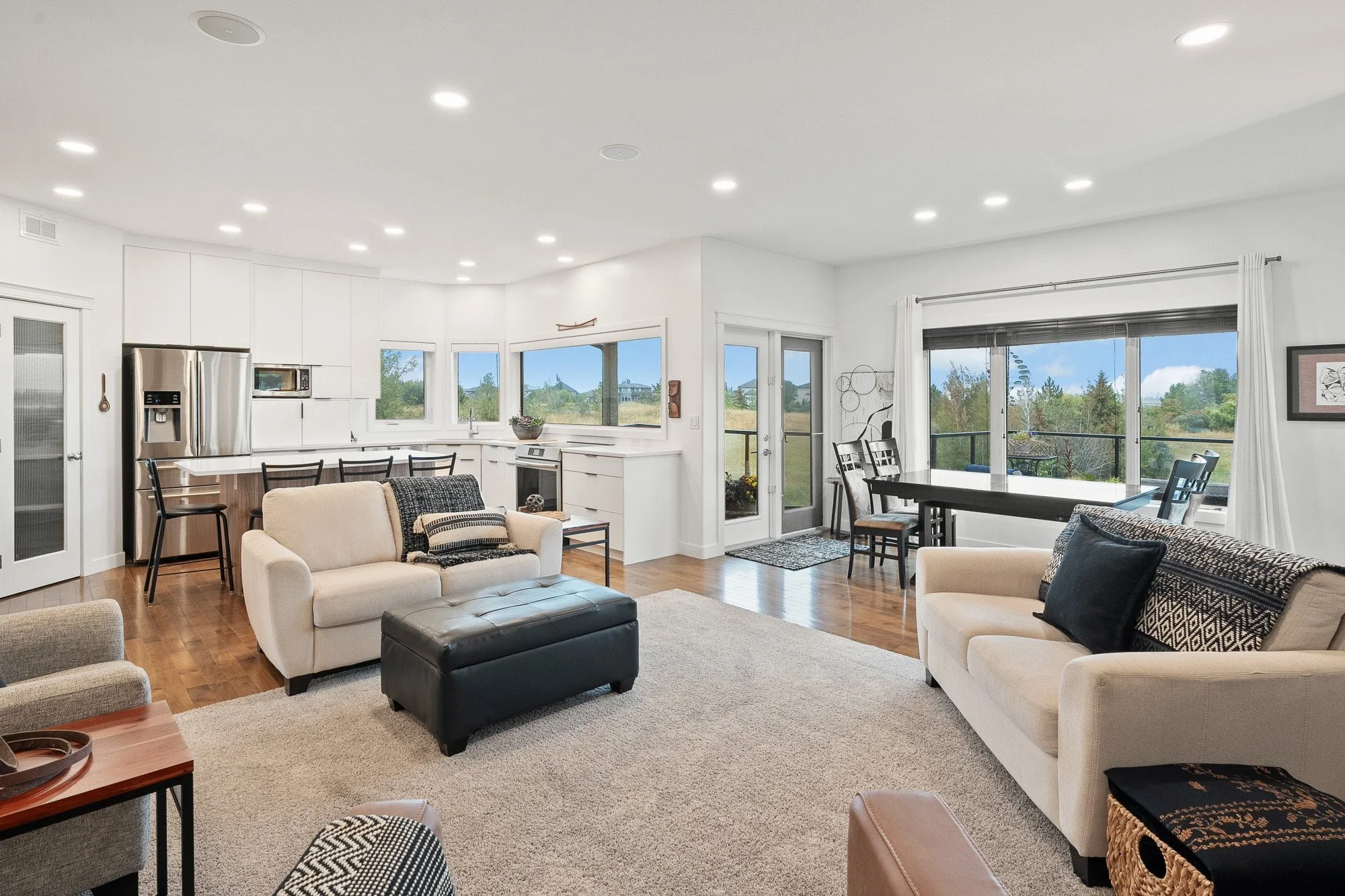 Modern open-concept living room and kitchen with neutral decor, large windows, and a view of greenery outside.