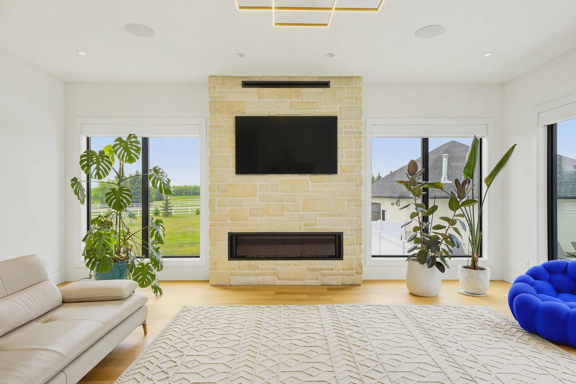 Sunlit living room featuring a stone fireplace, modern lighting and large windows overlooking the yard