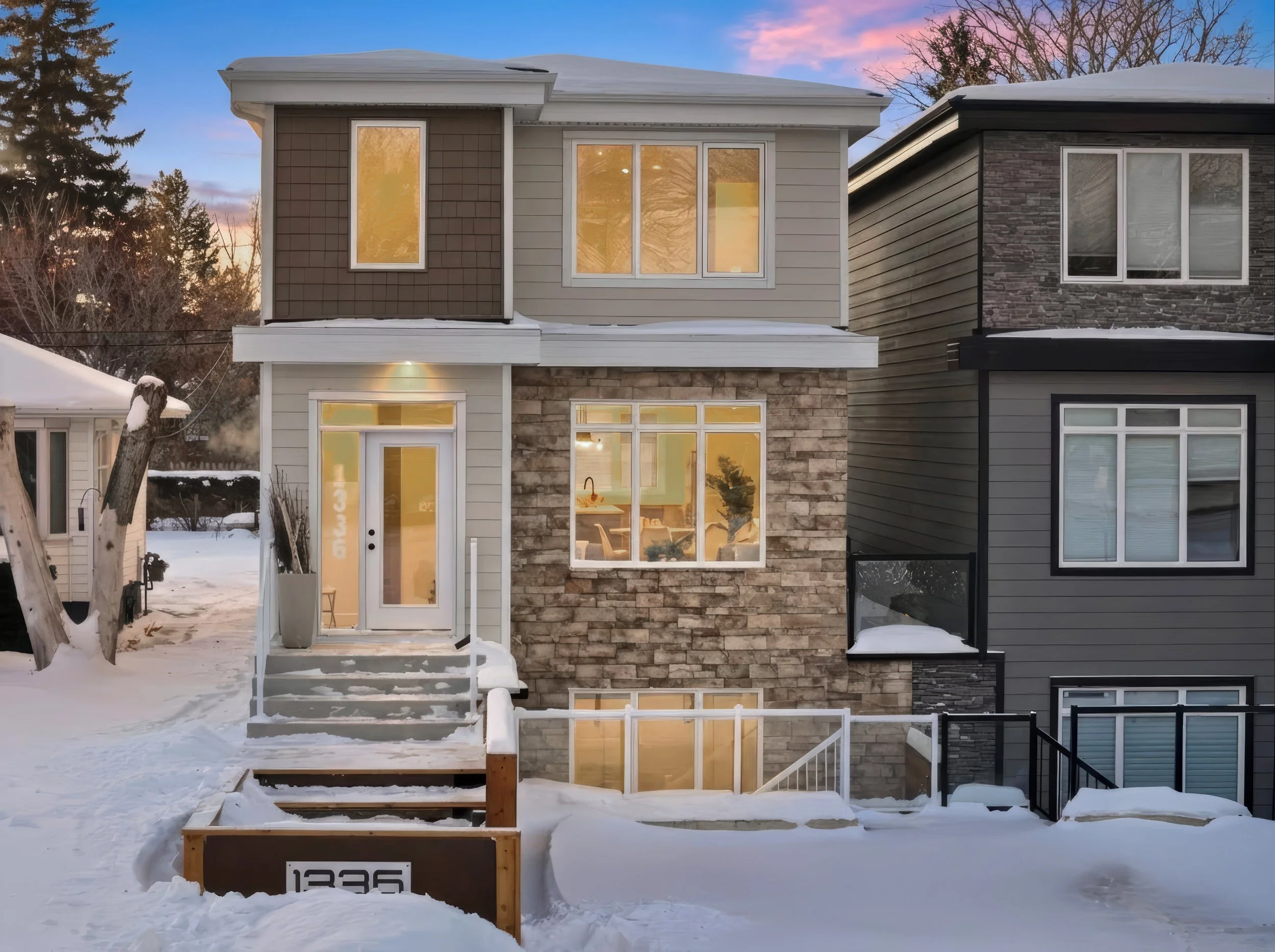 Modern two-story house with large windows and stone accents, surrounded by snow at sunset. Virtual twilight photography enhances the scene.