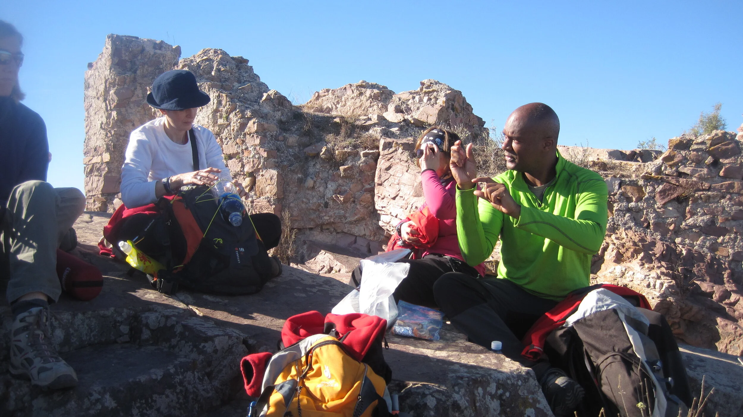 Group of hikers resting on rocky terrain with ruins in the background, some adjusting gear and talking.