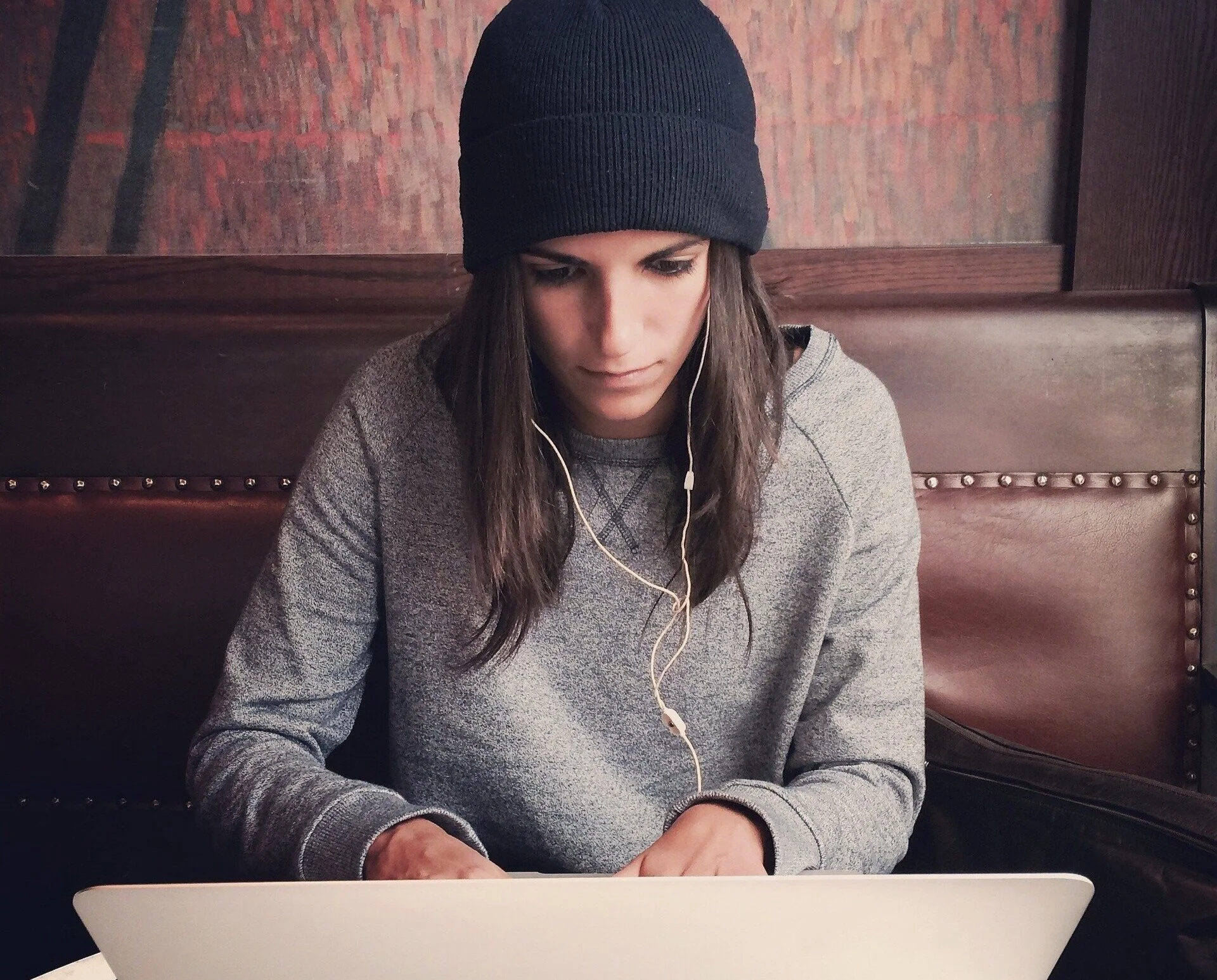 A woman wearing a black beanie hat and gray sweatshirt sitting at a table in a cafe or restaurant, looking at her laptop screen with earphones in her ears.