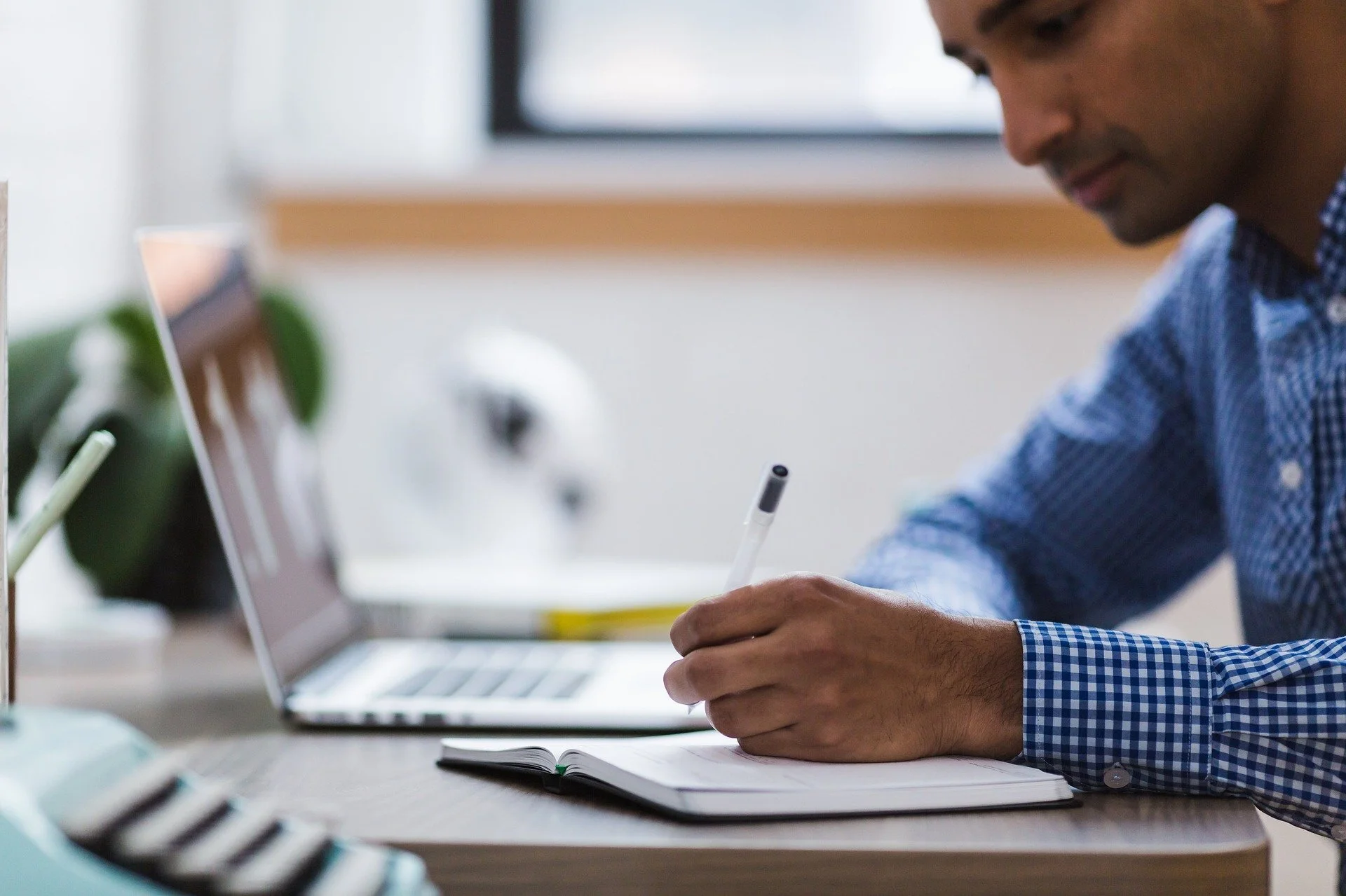 A man wearing a blue checkered shirt writing in an open notebook at a desk, with a laptop, a plant, and other office items in the background.
