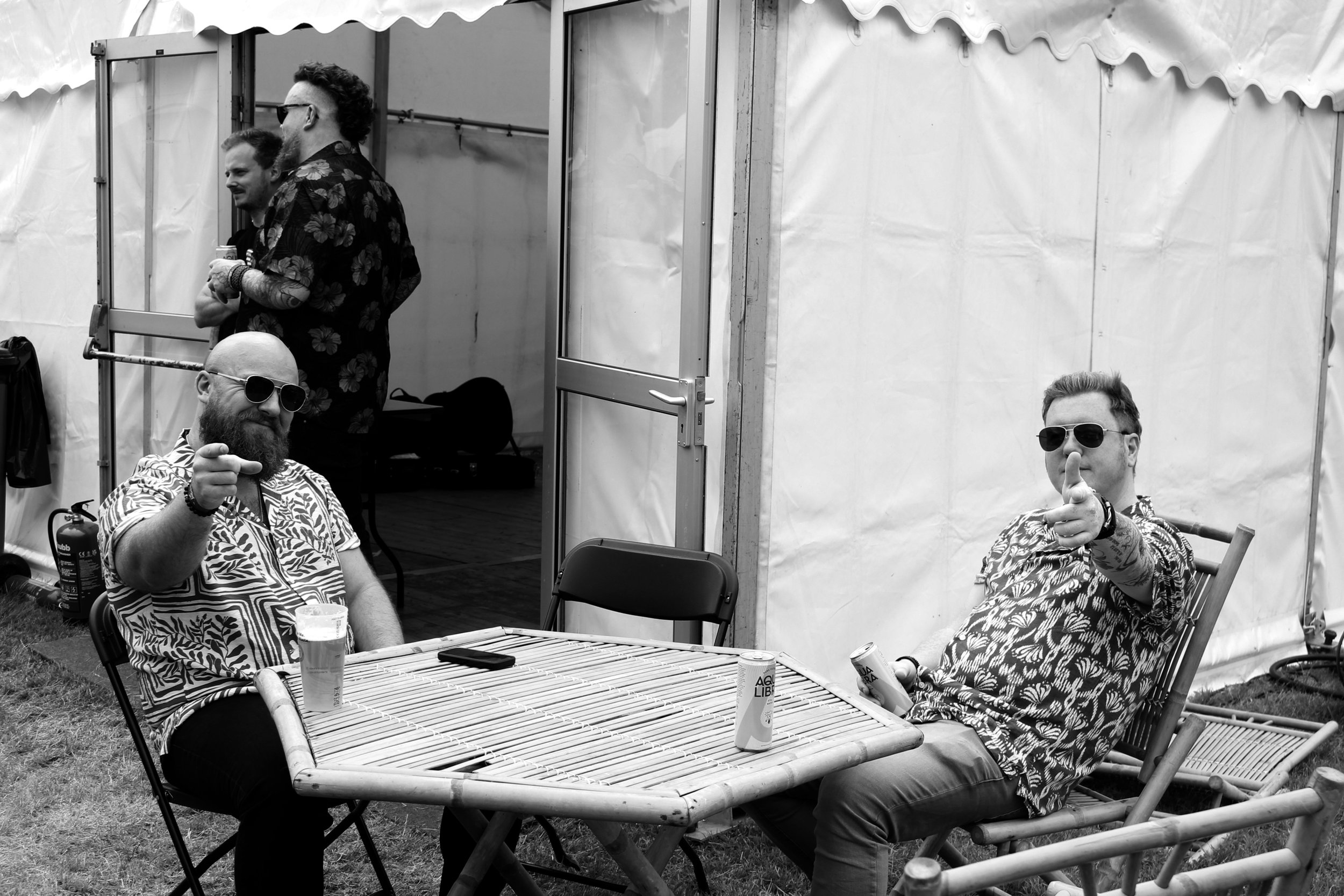 Four men with sunglasses, two sitting at a bamboo outdoor table, two standing behind, at a casual gathering under a tent with white walls and a scalloped roof. One man is making a pointing gesture toward the camera, another is holding a drink.
