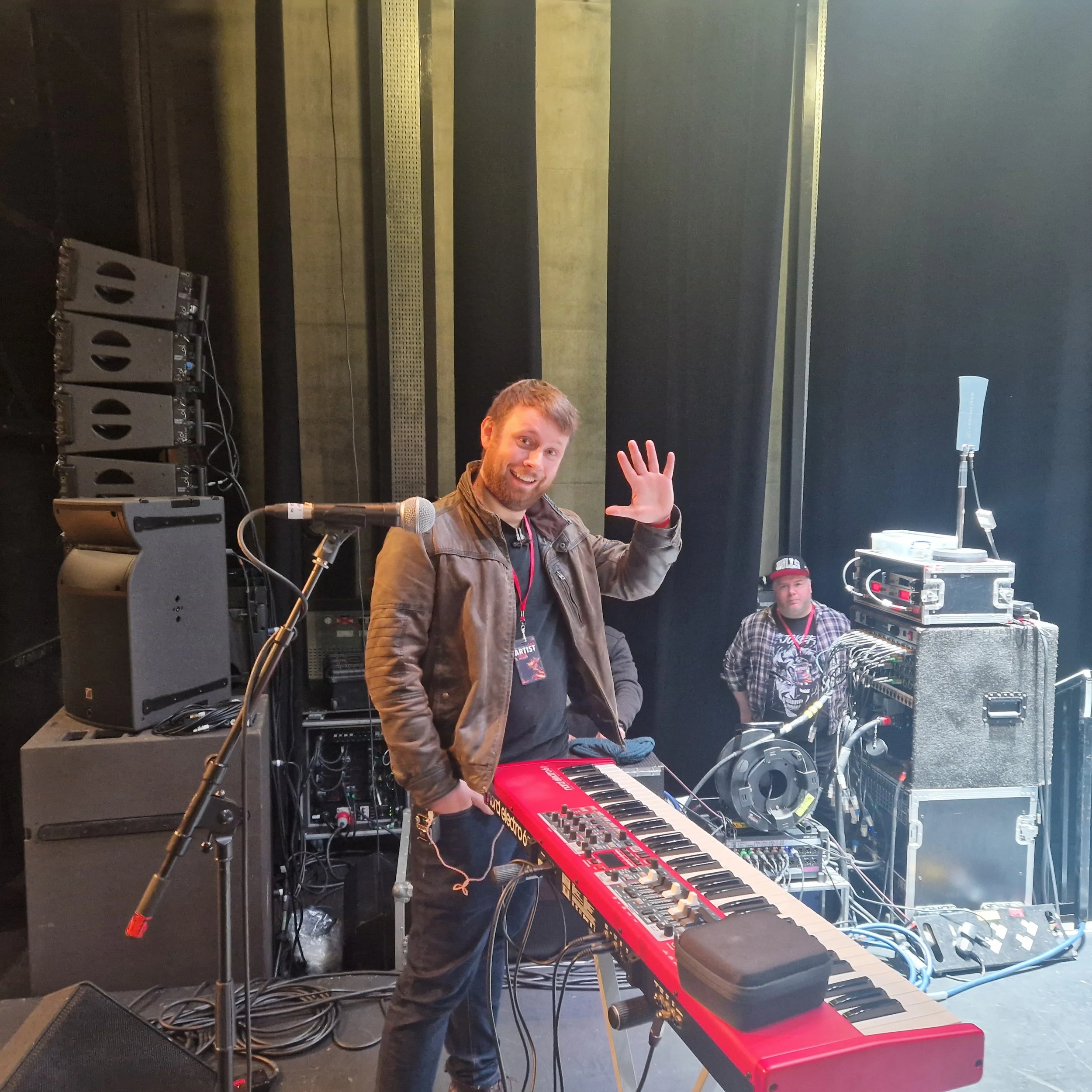A man with short hair and a beard waving while standing behind a red electronic keyboard on a stand. In the background, there is sound equipment, and another person in a plaid shirt is visible. The setting appears to be a stage with black walls and professional audio gear.